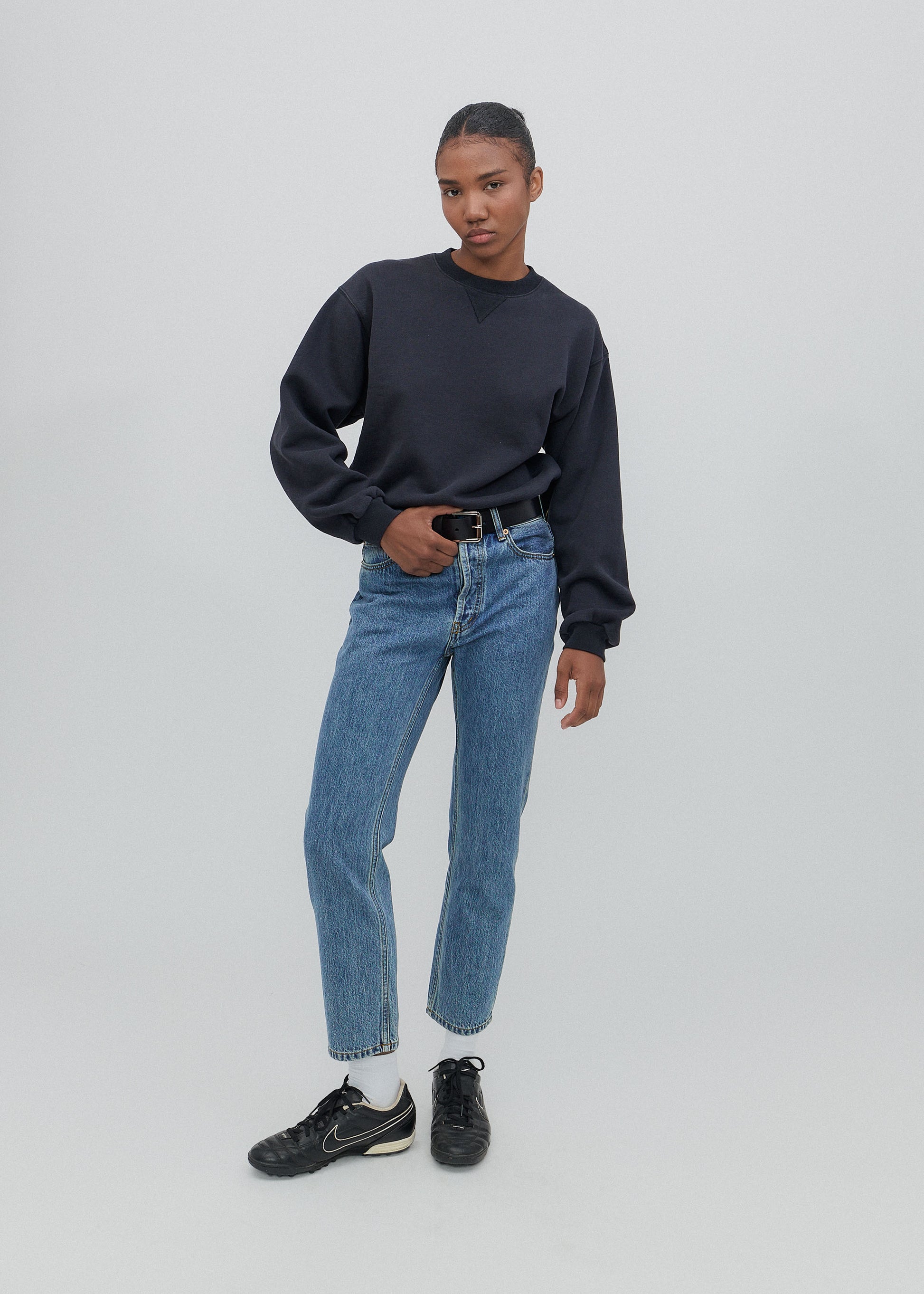 Person wearing a black sweater, blue jeans, and black sneakers on a light gray background