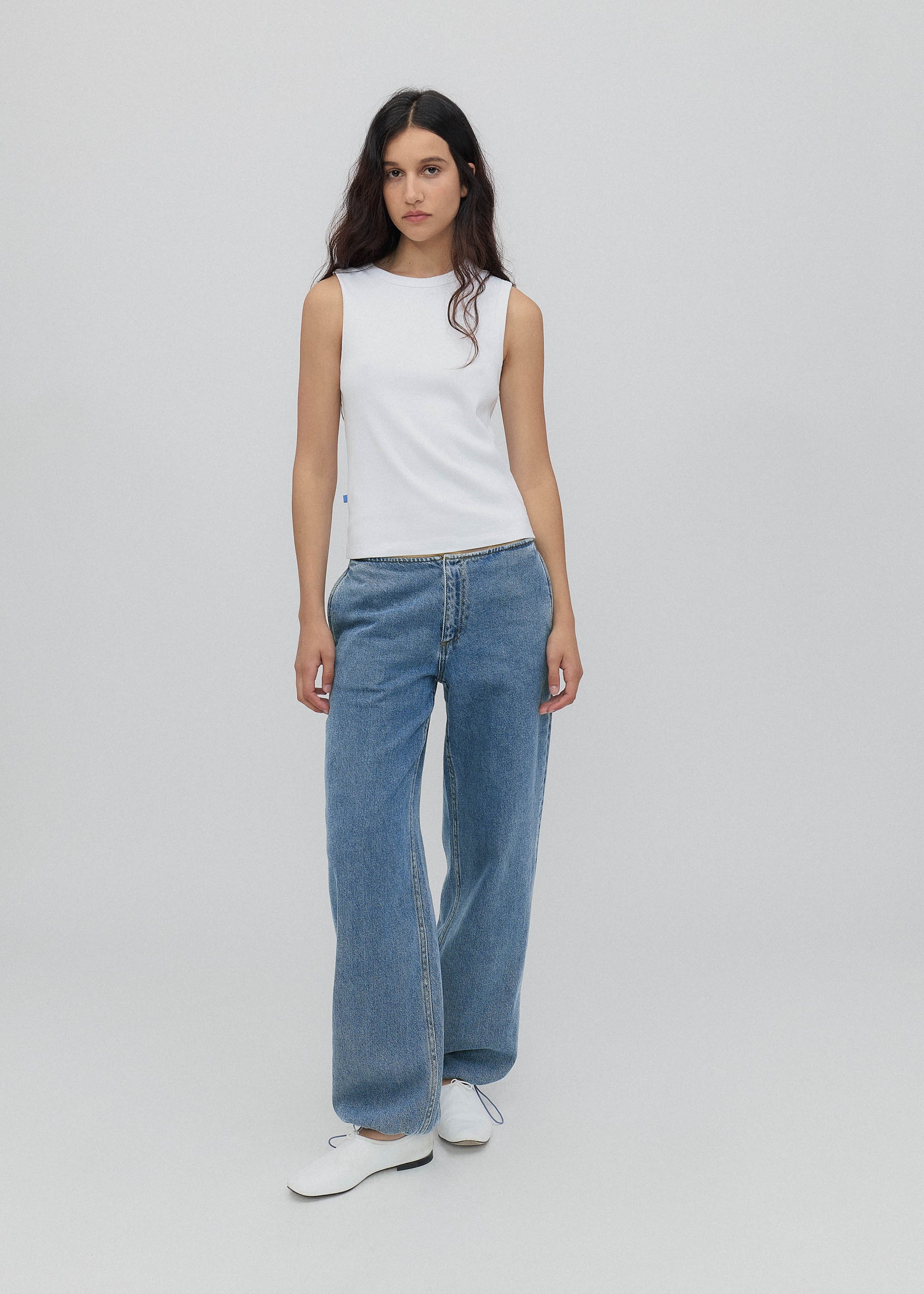 Woman wearing a white sleeveless top and blue jeans on a plain background
