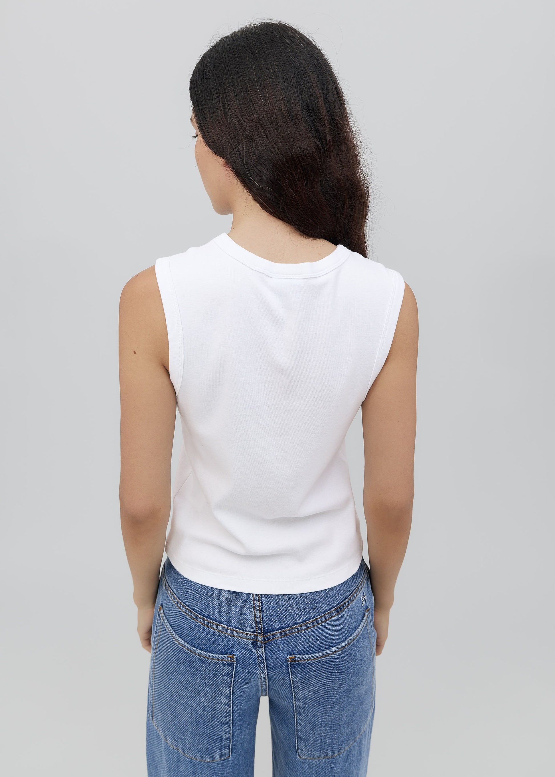 Person wearing a white sleeveless top and blue jeans on a plain background