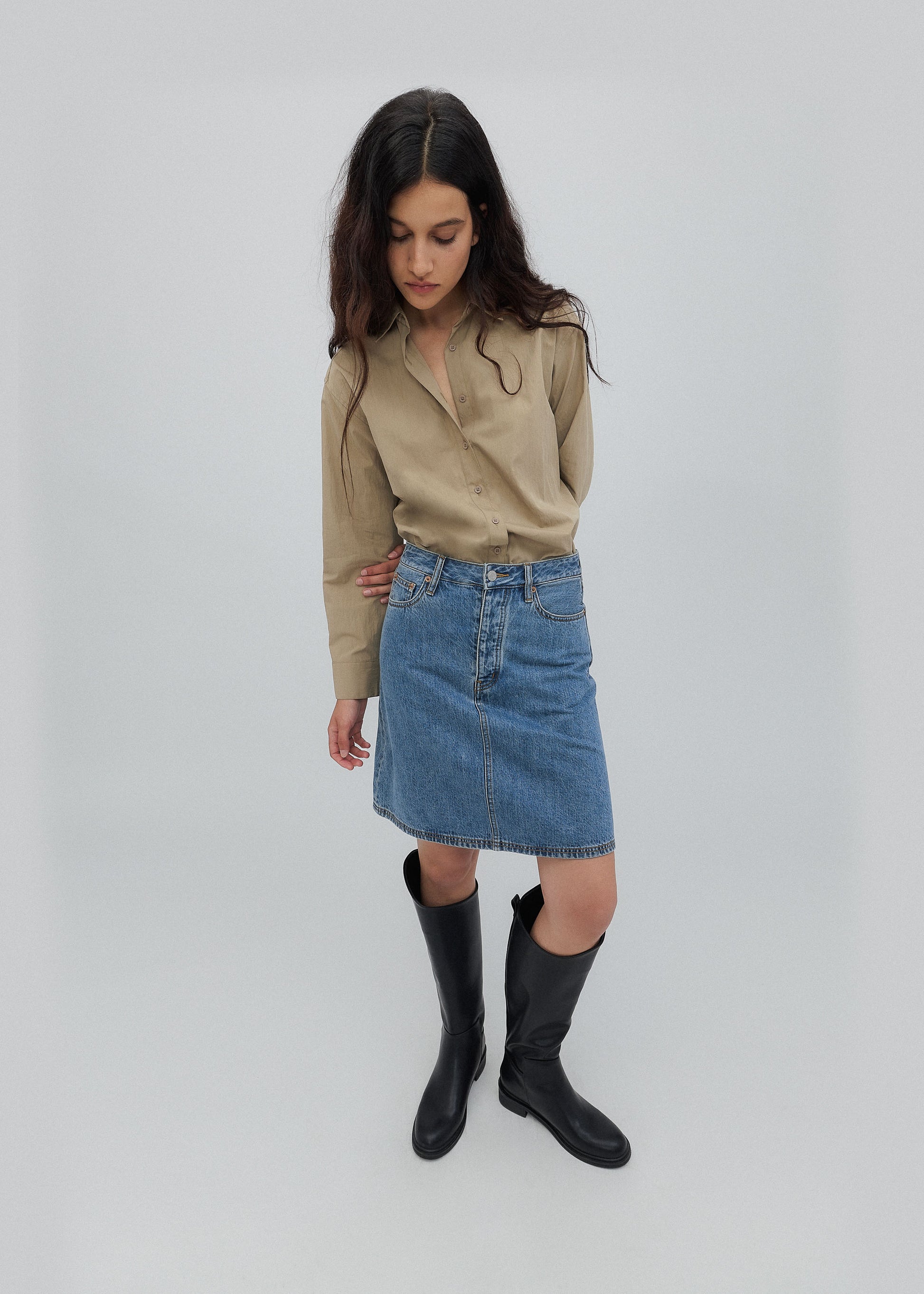 Woman wearing a beige blouse, blue denim skirt, and black boots on a white background