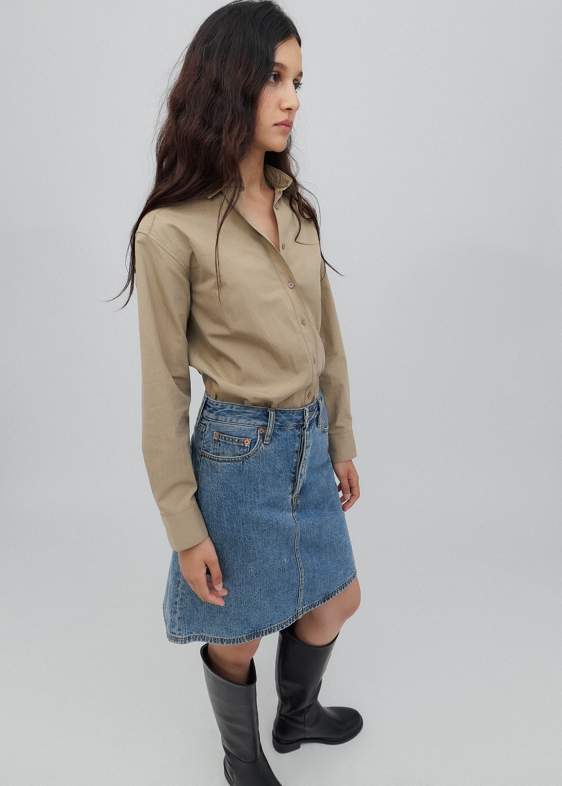 Woman wearing a beige shirt, blue denim skirt, and black rain boots on a plain background