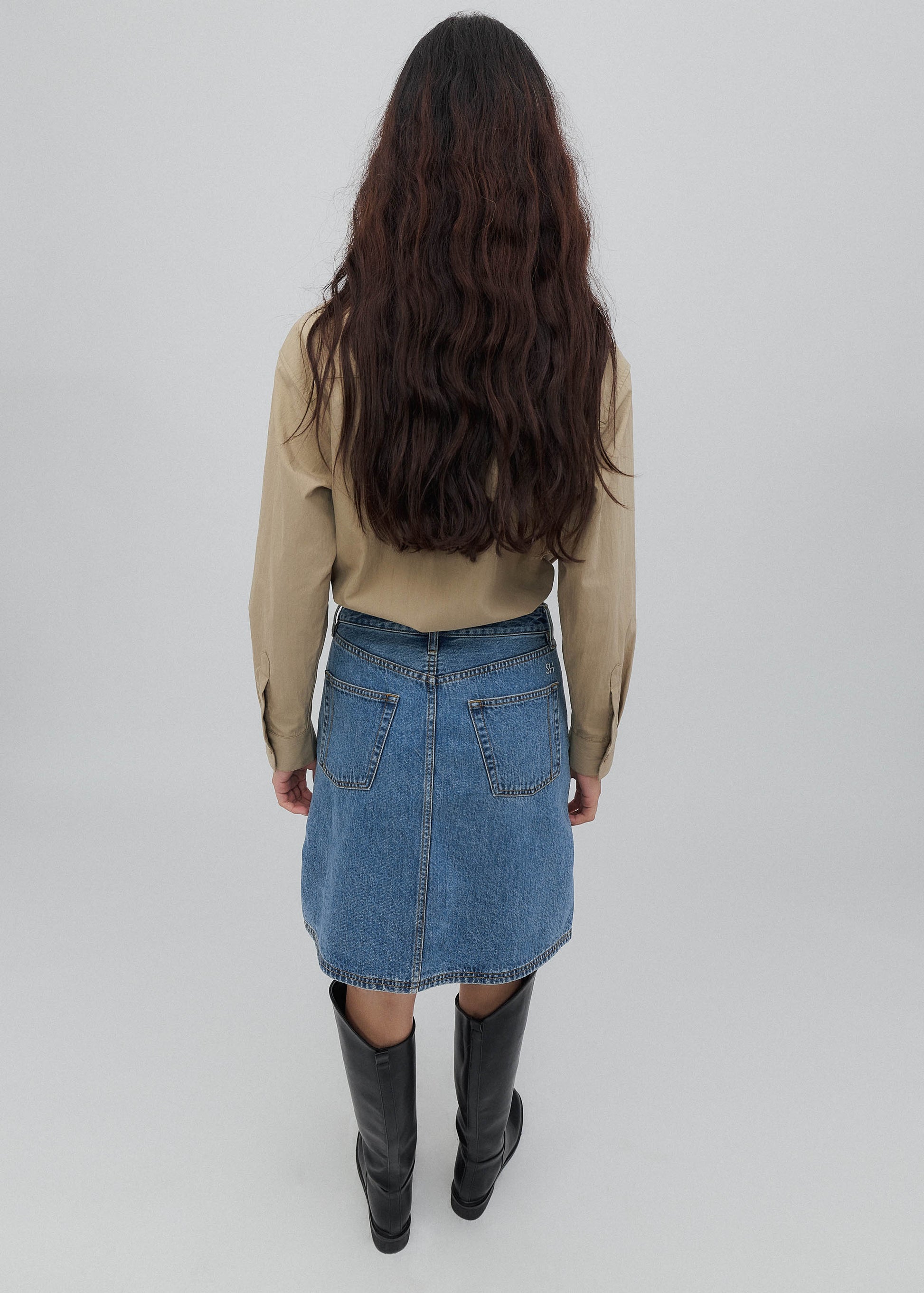 Person wearing a beige jacket, blue denim skirt, and black boots on a white background