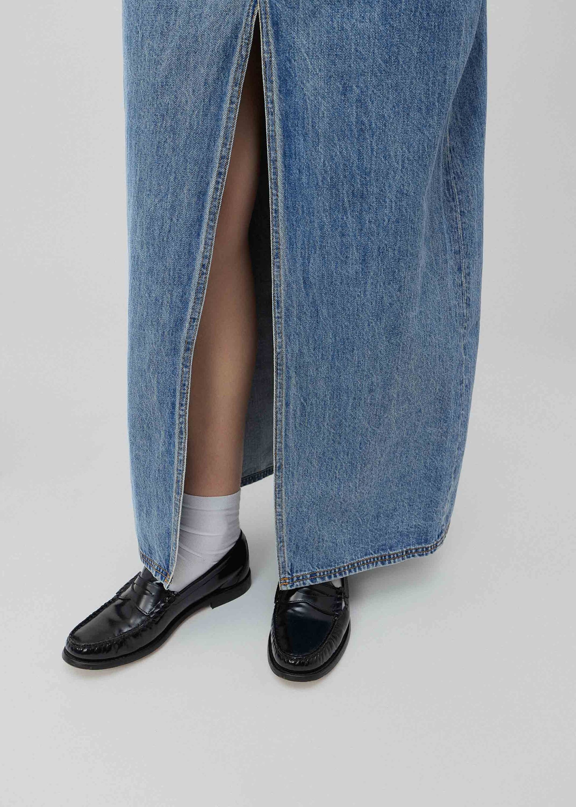 Person wearing a long blue denim skirt with black shoes and white socks on a light gray background