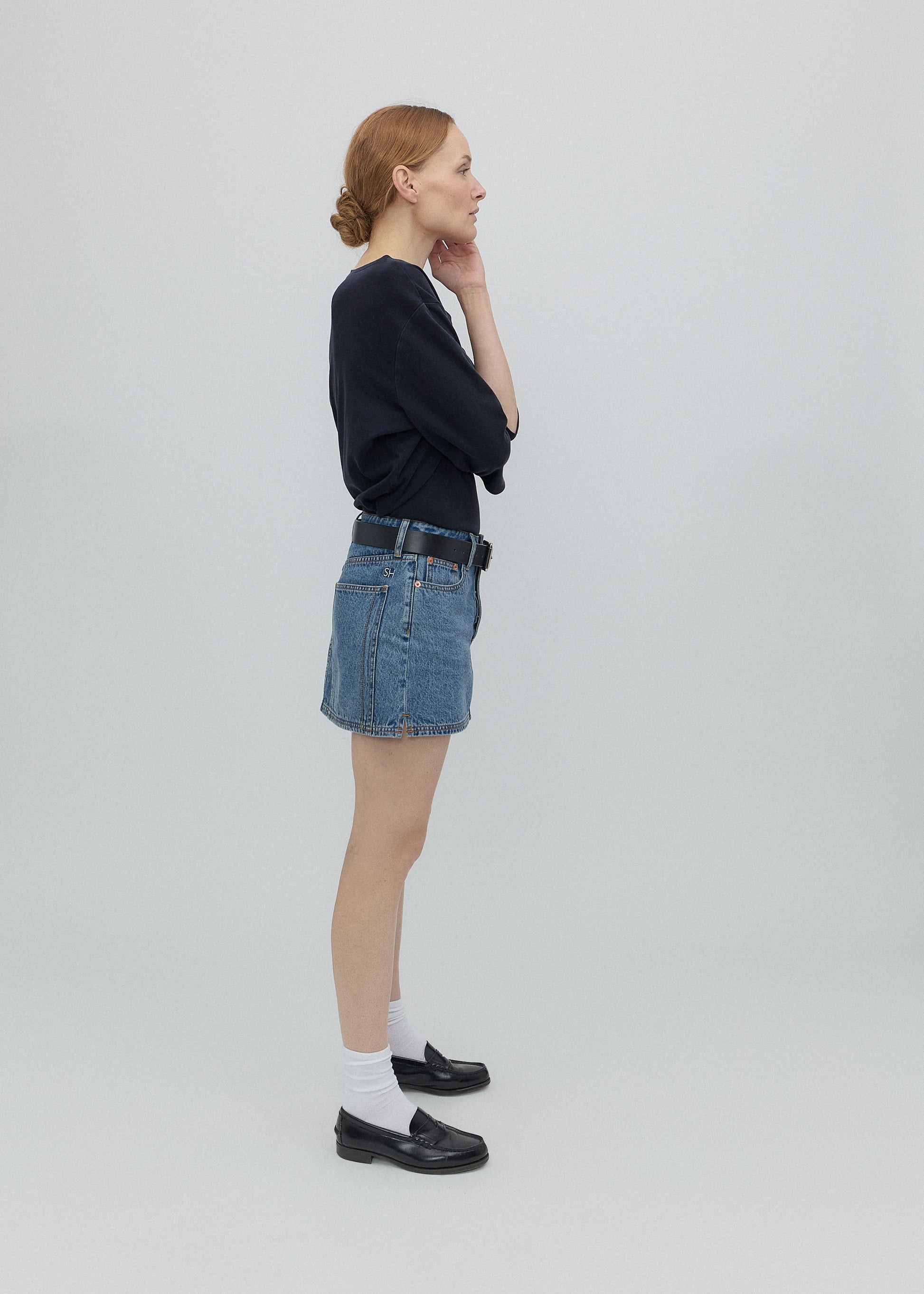 Woman wearing a black sweater, blue denim skirt, and white socks with black shoes on a light gray background