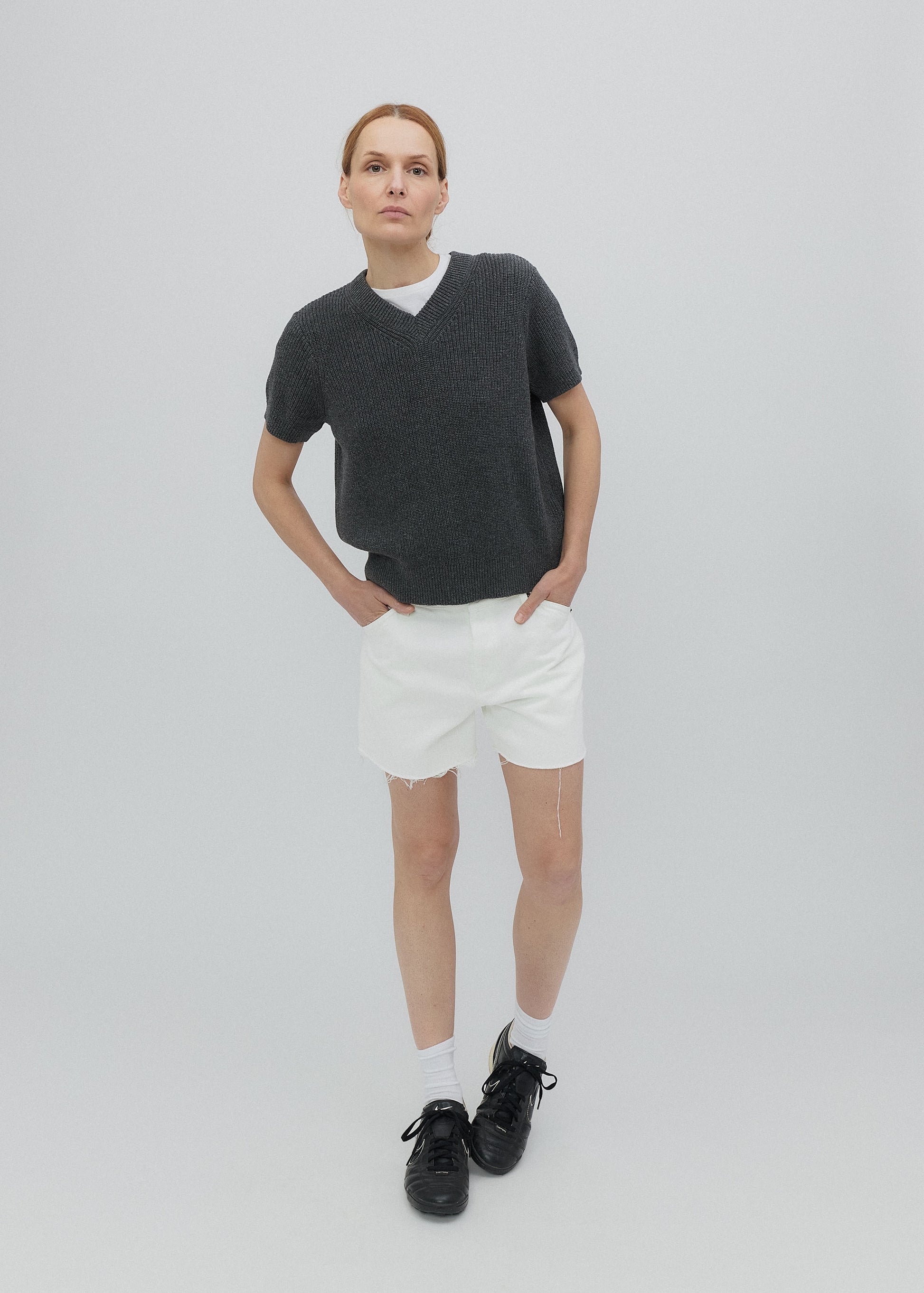 Person wearing a dark gray V-neck sweater, white shorts, and black shoes on a light gray background
