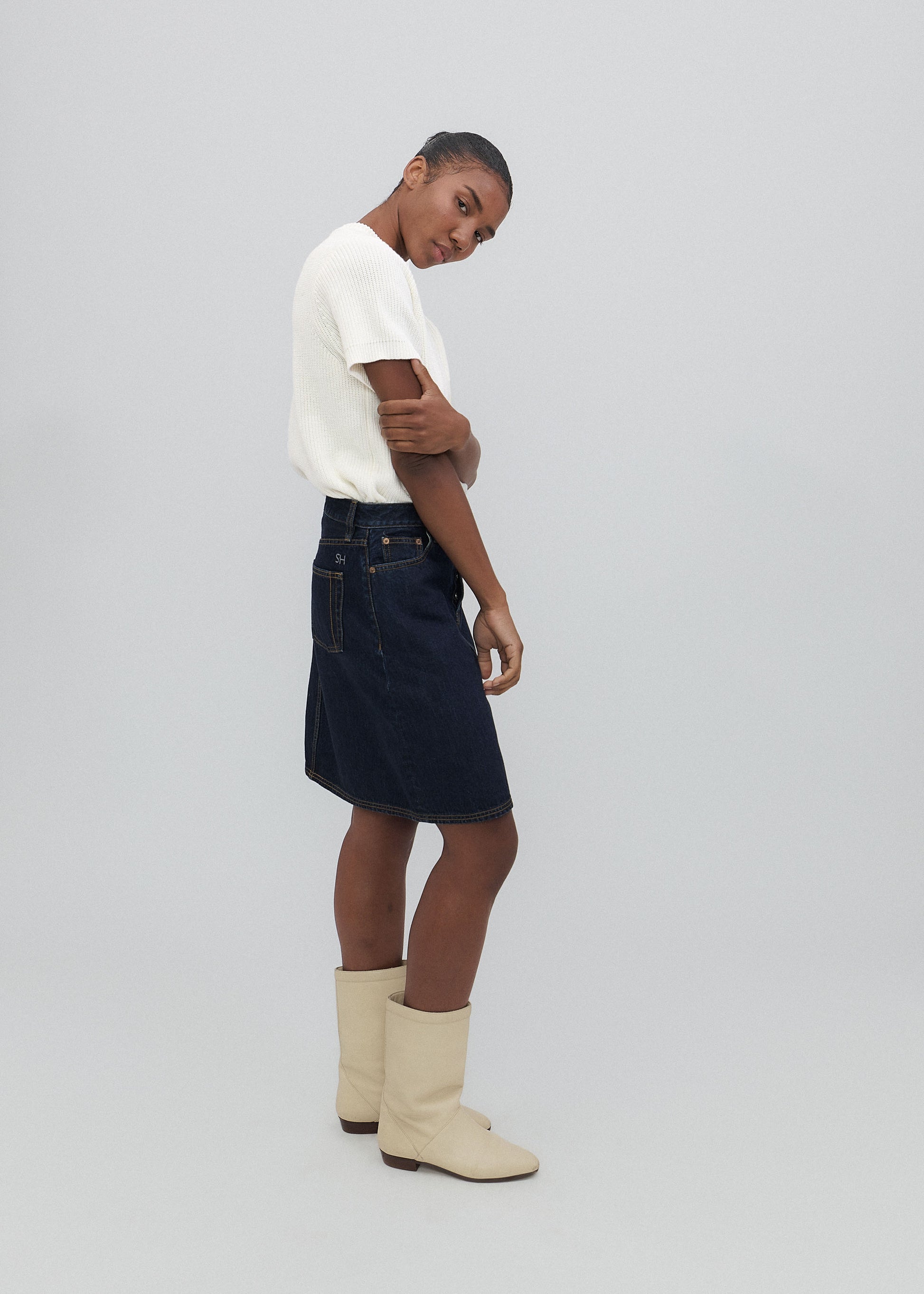 Person wearing a white t-shirt, black denim skirt, and beige boots on a light gray background
