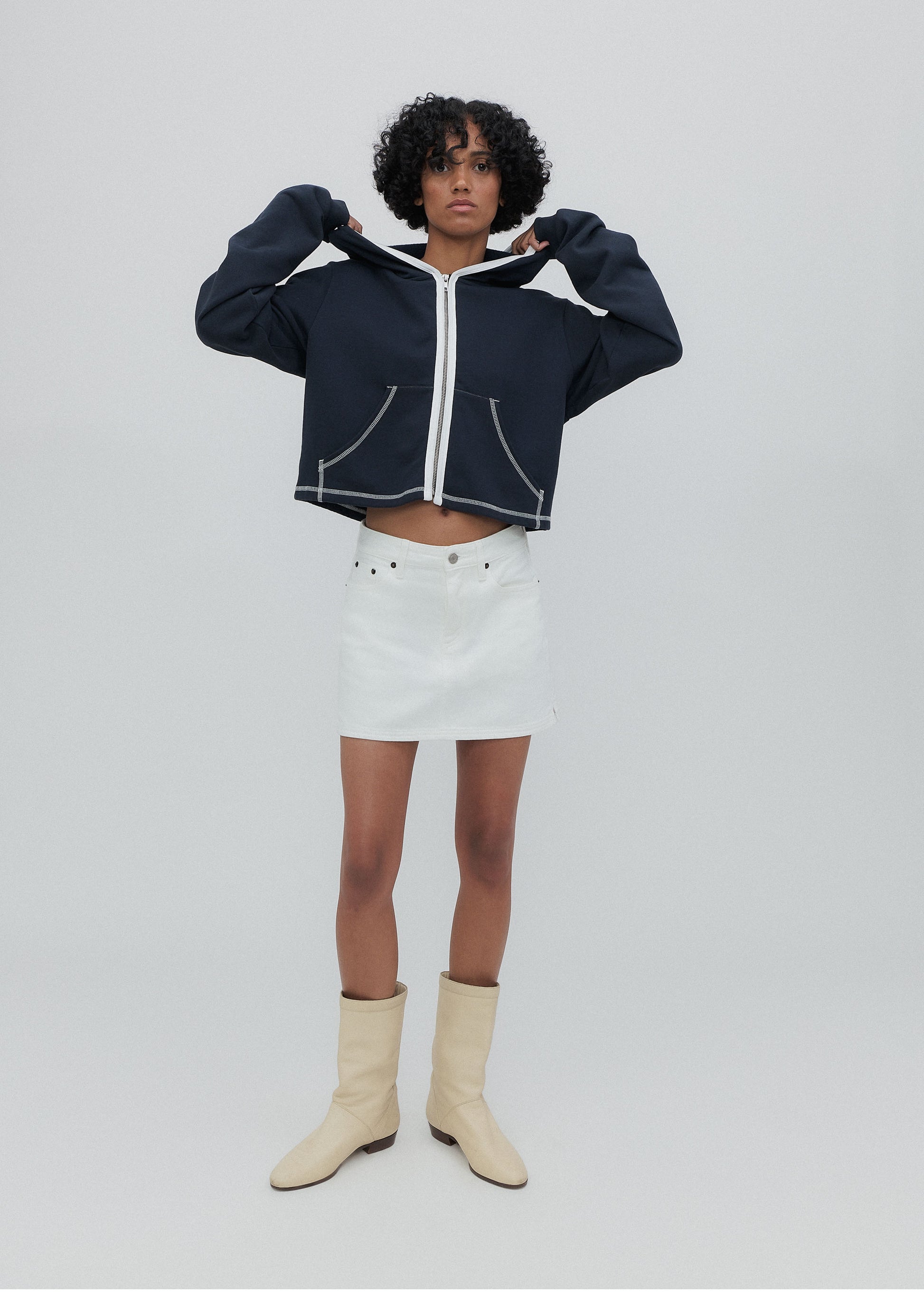 Person wearing a navy blue hoodie, white skirt, and beige boots on a light gray background