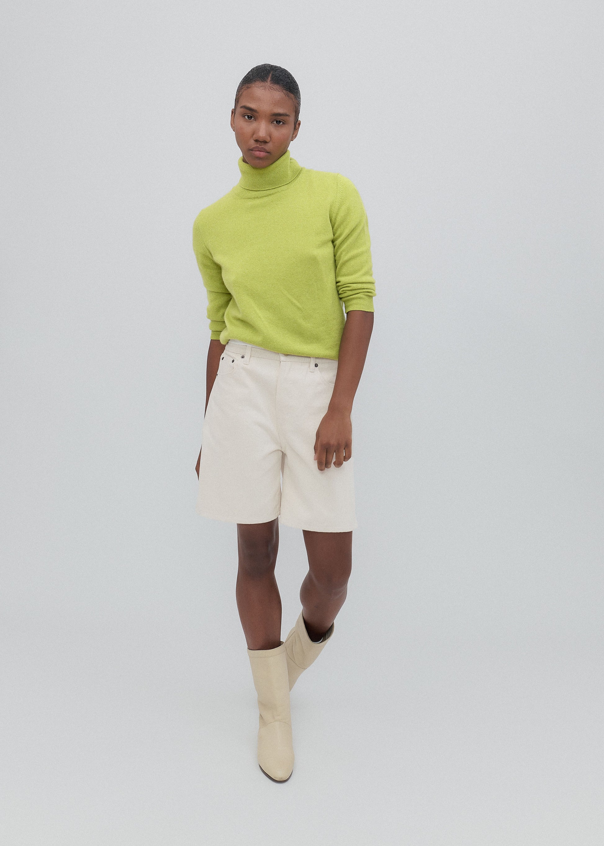 Person wearing a bright green sweater, white shorts, and beige boots on a plain background