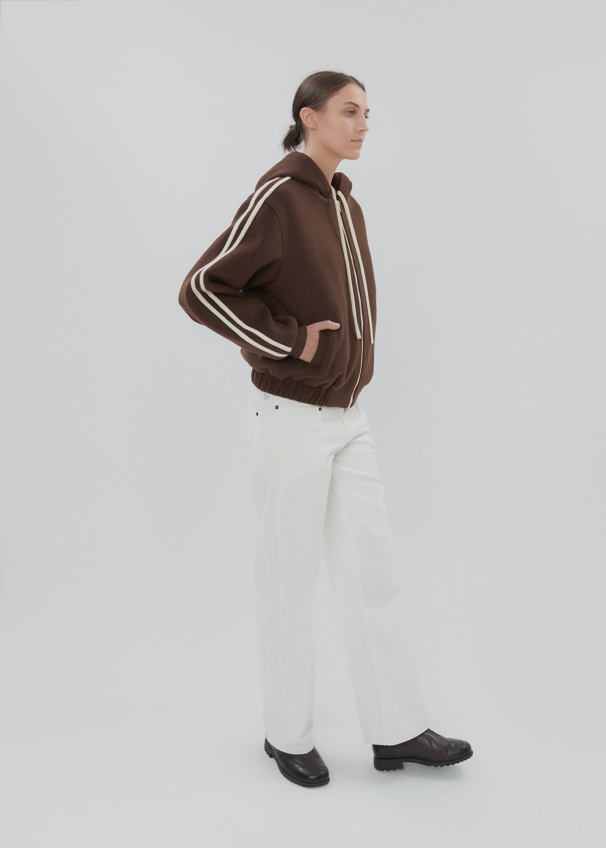 Person wearing a brown jacket with white stripes and white pants on a light gray background