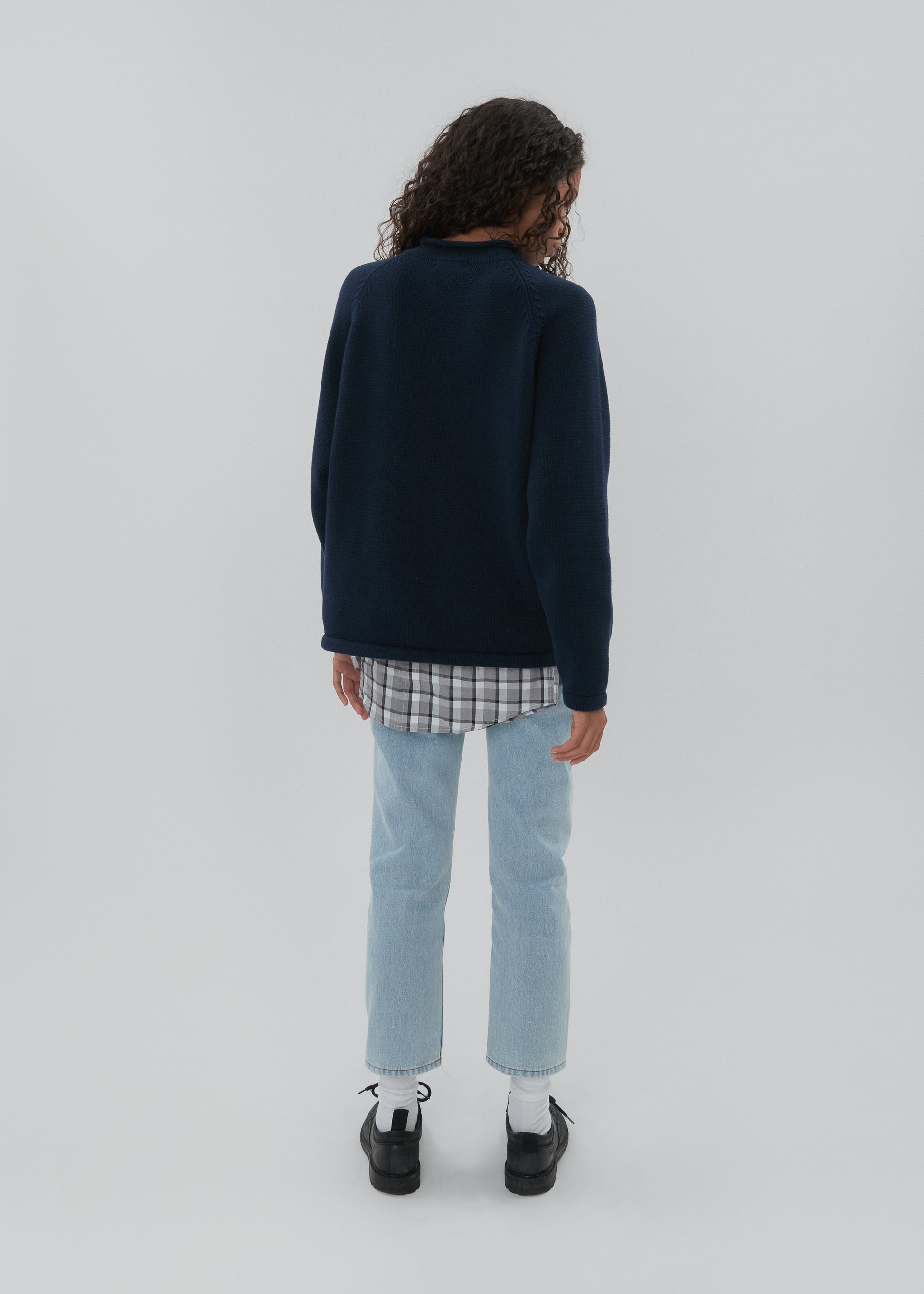 Person wearing a navy sweater, plaid shirt, light blue jeans, and black shoes on a white background