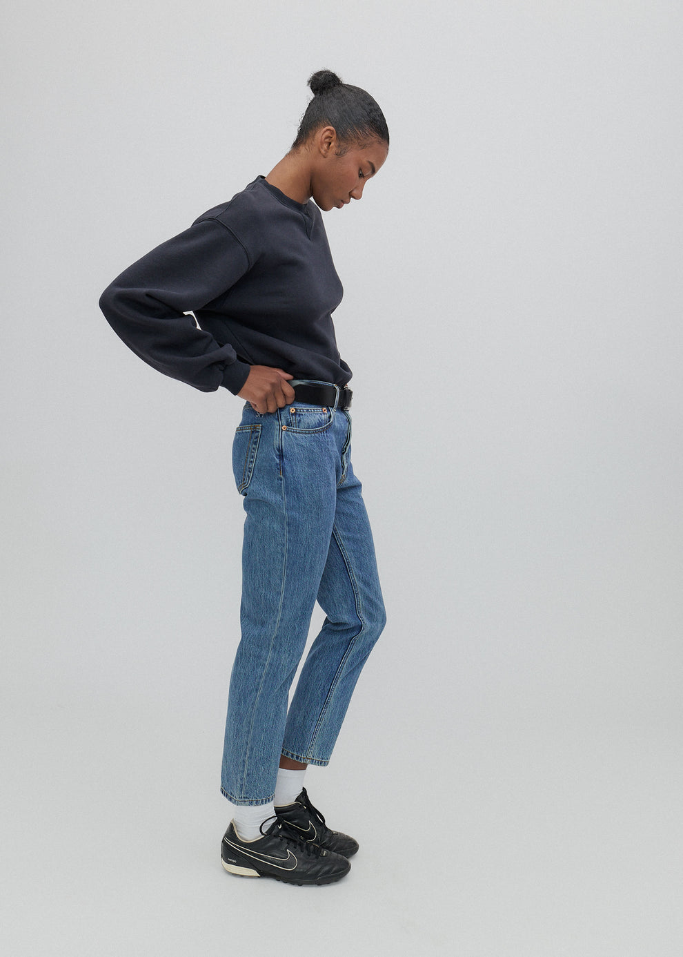 Person wearing a navy sweatshirt, blue jeans, and black sneakers on a light gray background