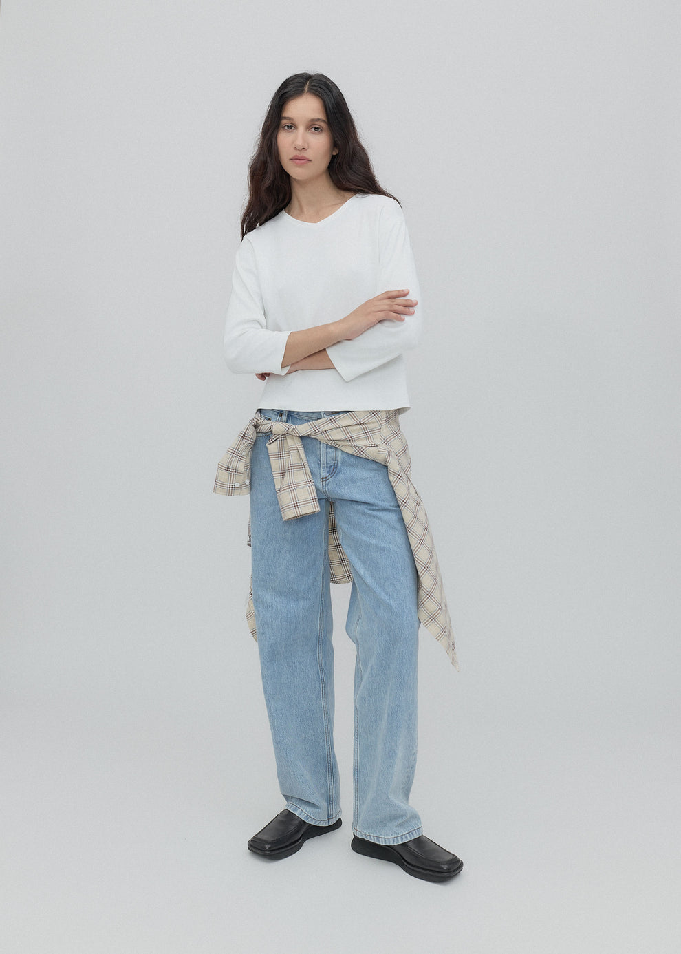 Woman wearing a white sweater, light blue jeans, and black shoes on a plain background