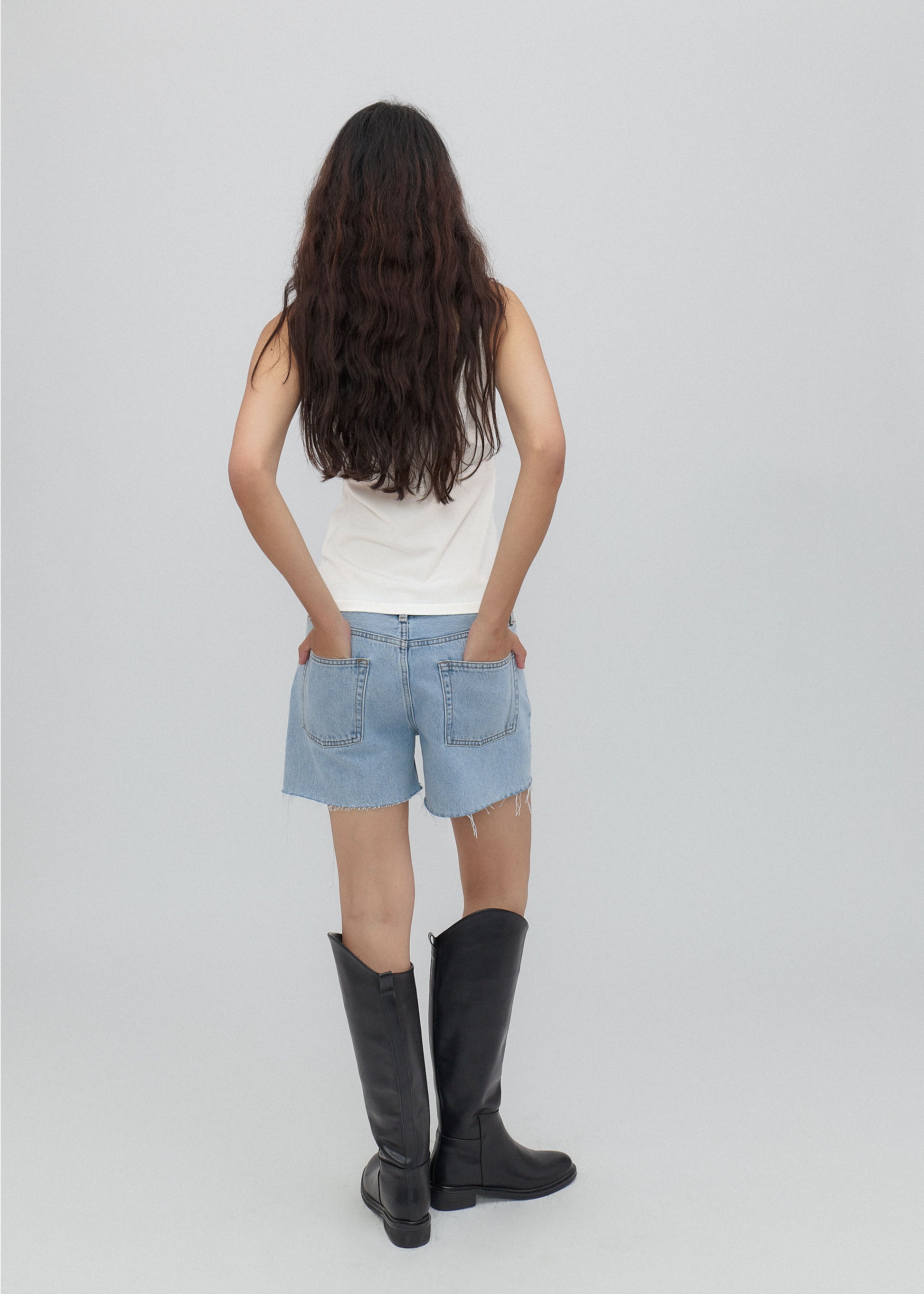 Person wearing a white tank top, light blue denim shorts, and black rain boots on a plain background