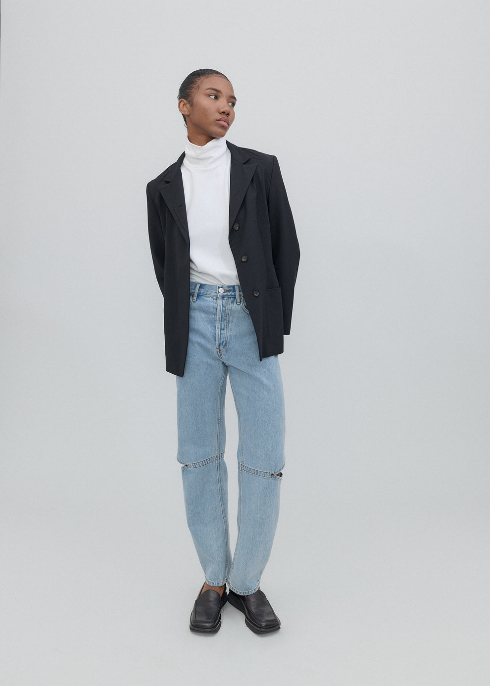 Person wearing a black blazer, white turtleneck, light blue jeans, and black shoes on a plain background