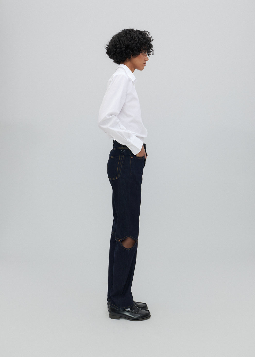 Person wearing a white shirt and dark jeans with a hole, standing against a plain background