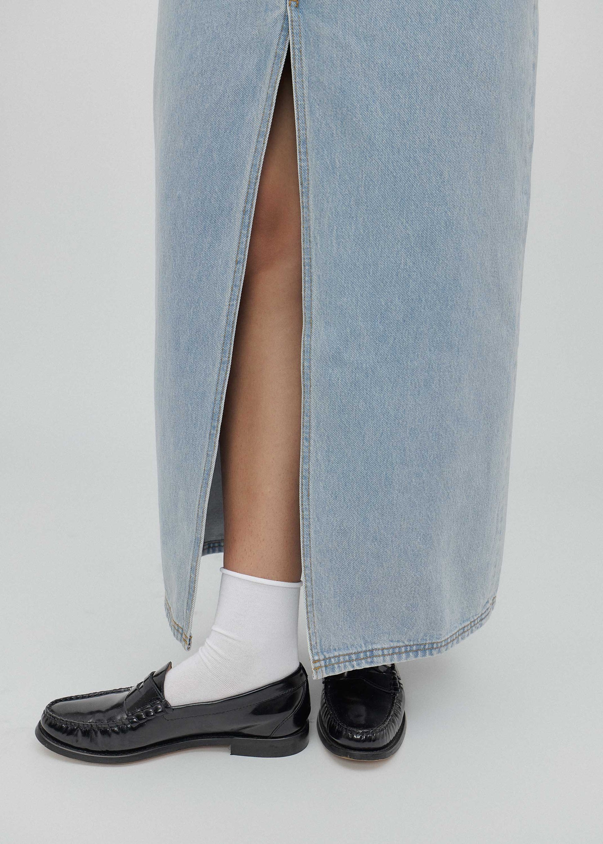 Person wearing a long denim skirt with black shoes and white socks on a light gray background