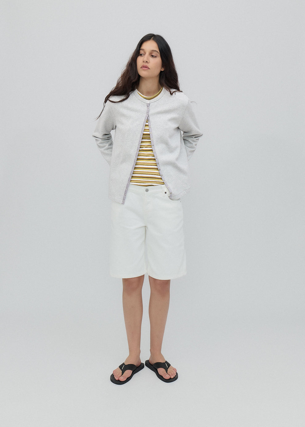 Woman wearing a light gray jacket, striped shirt, and white shorts on a plain background