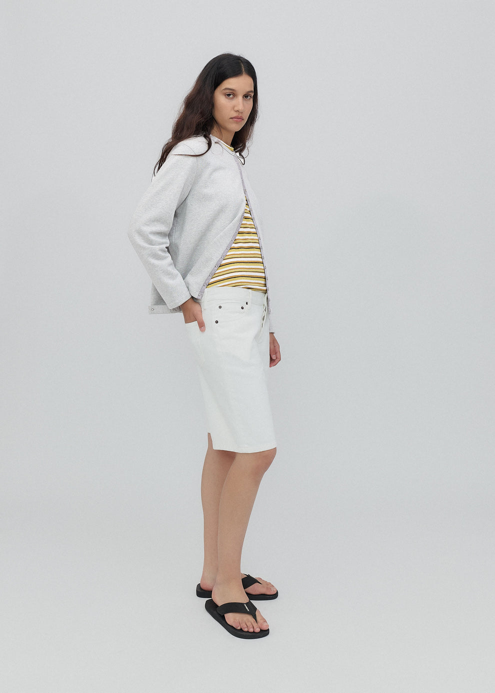 Woman wearing a light gray jacket, striped top, white skirt, and black sandals on a plain background