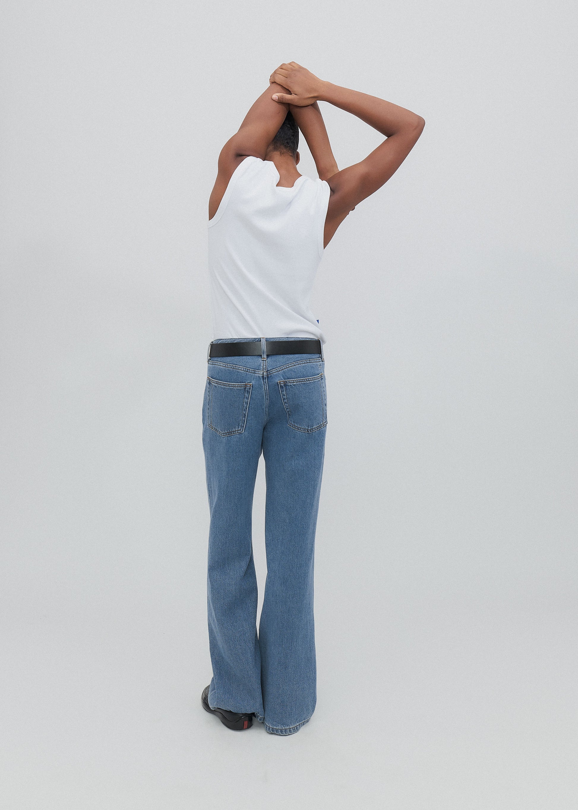 Person wearing a white shirt and blue jeans standing with hands on head against a light gray background