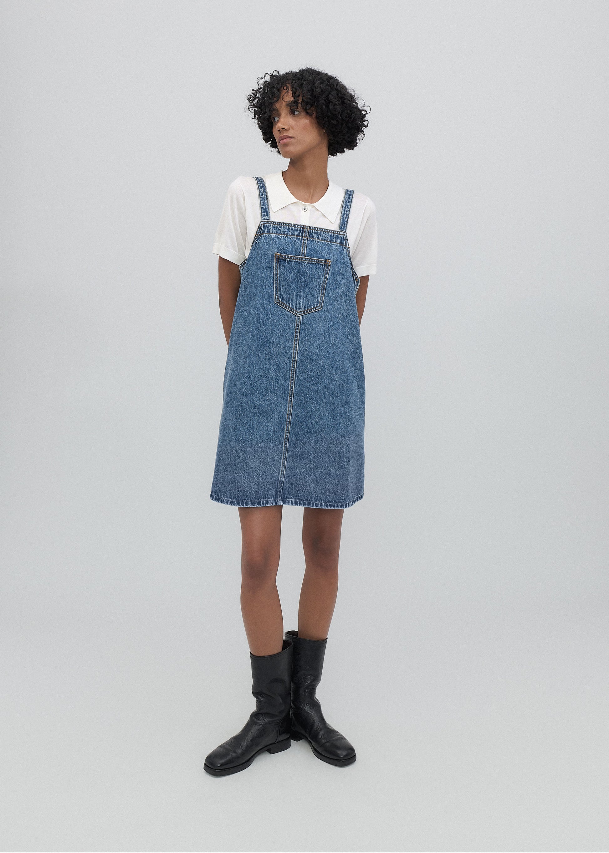 Person wearing a denim pinafore dress with a white shirt underneath on a plain background