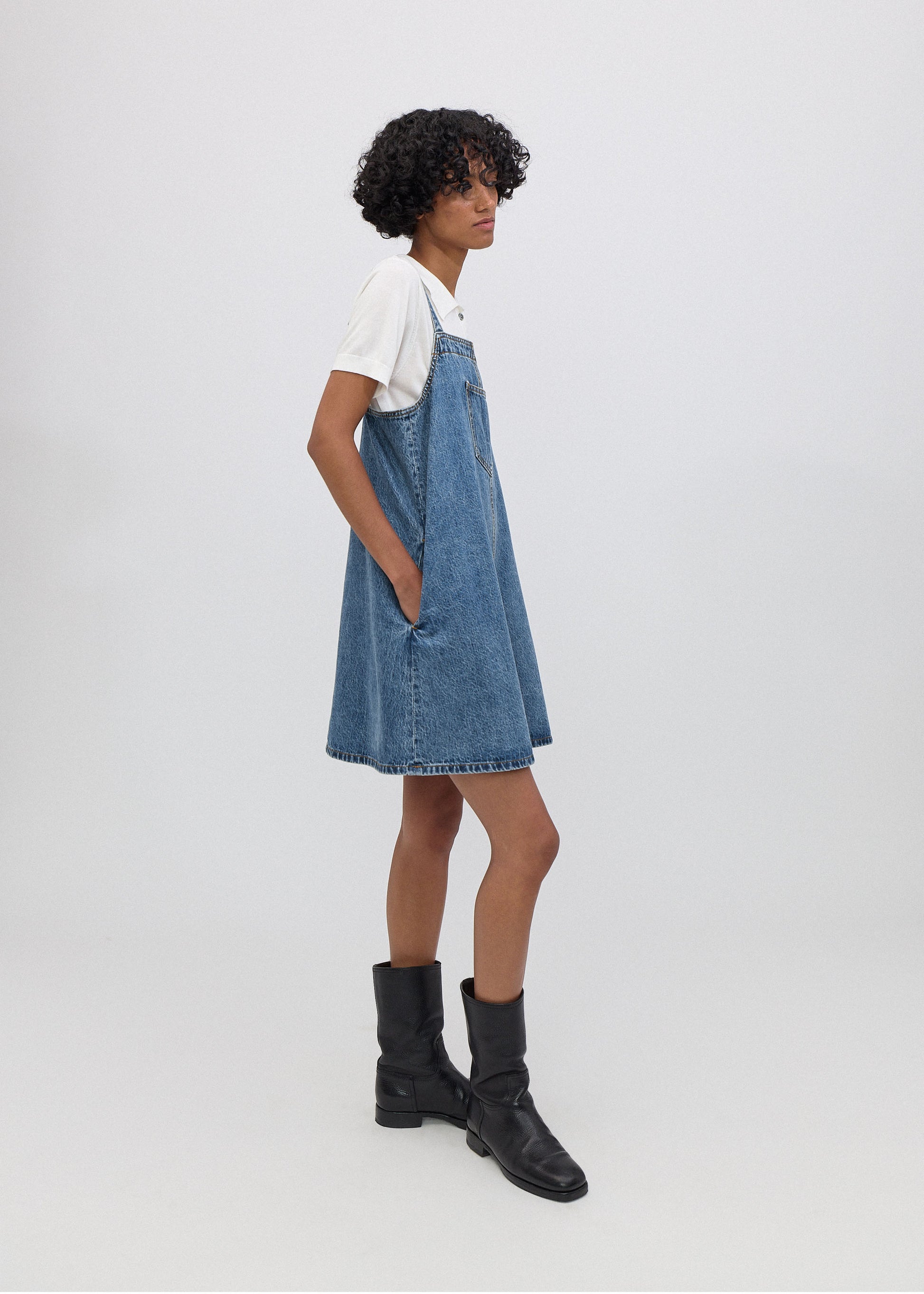 Person wearing a denim pinafore dress with a white shirt underneath on a plain background