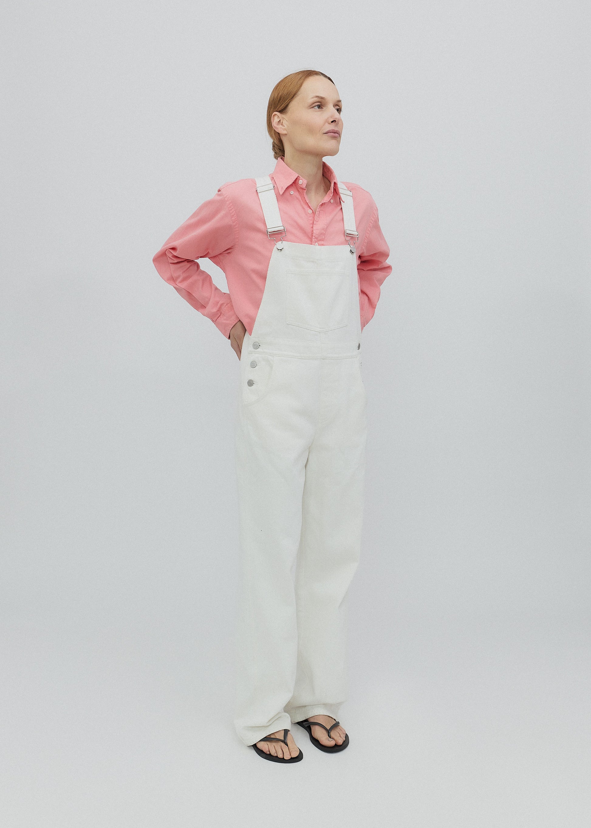 Woman wearing a pink shirt and white overalls on a light gray background