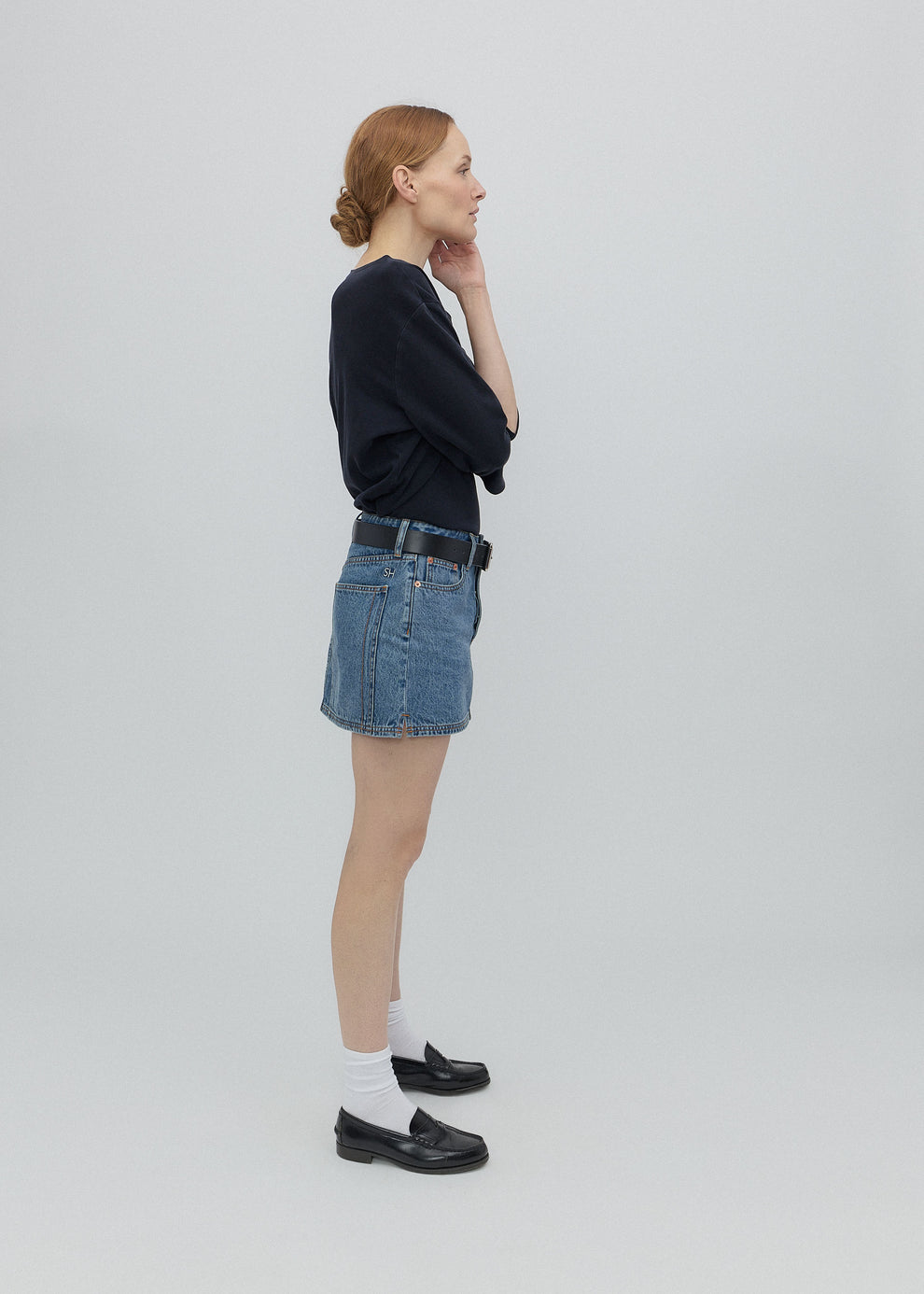 Woman wearing a black sweater, blue denim skirt, and white socks with black shoes on a light gray background