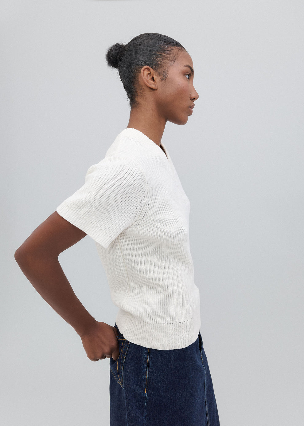 Woman wearing a white ribbed sweater and dark blue jeans against a light gray background