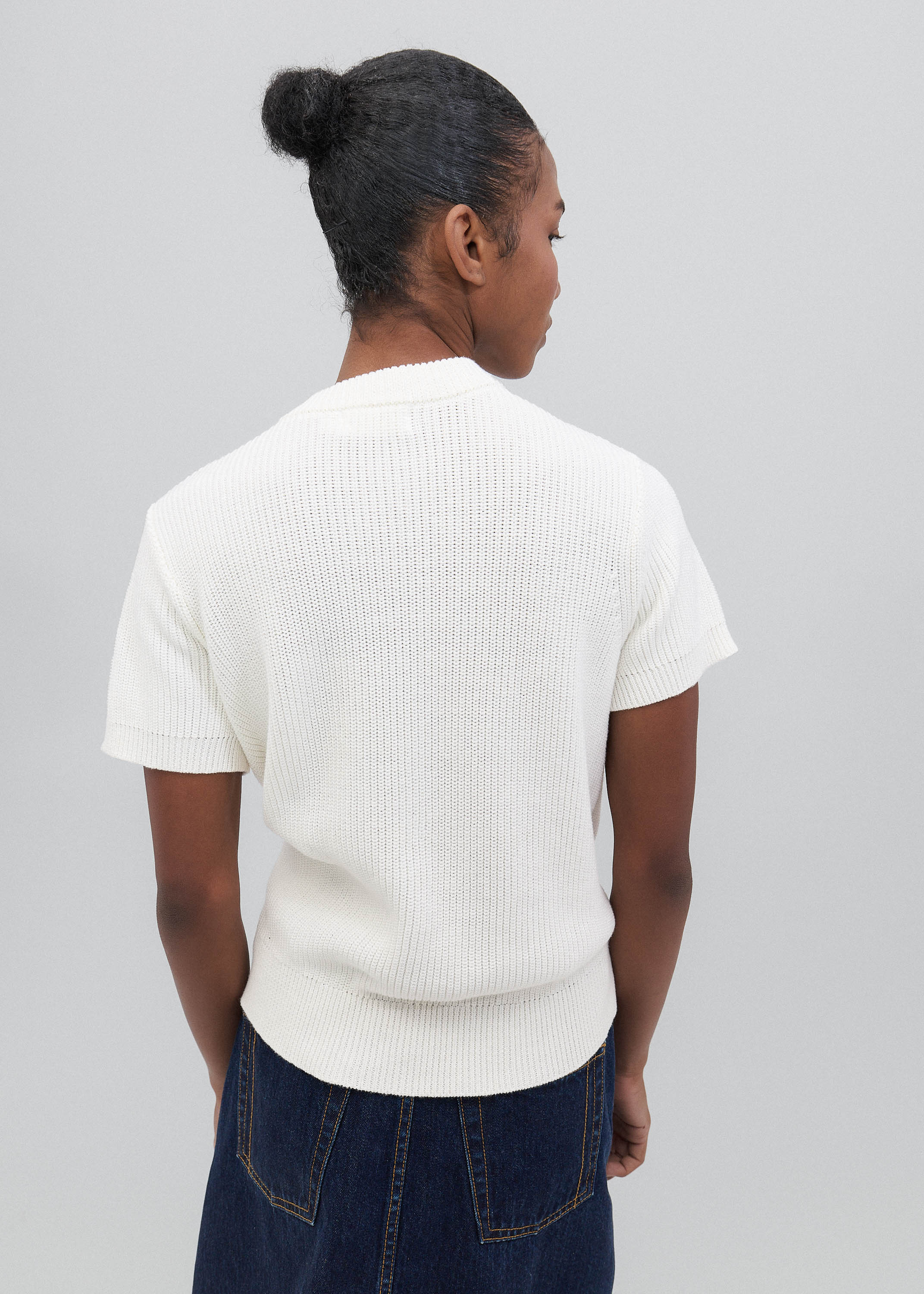 Person wearing a white ribbed sweater and dark blue jeans on a plain background
