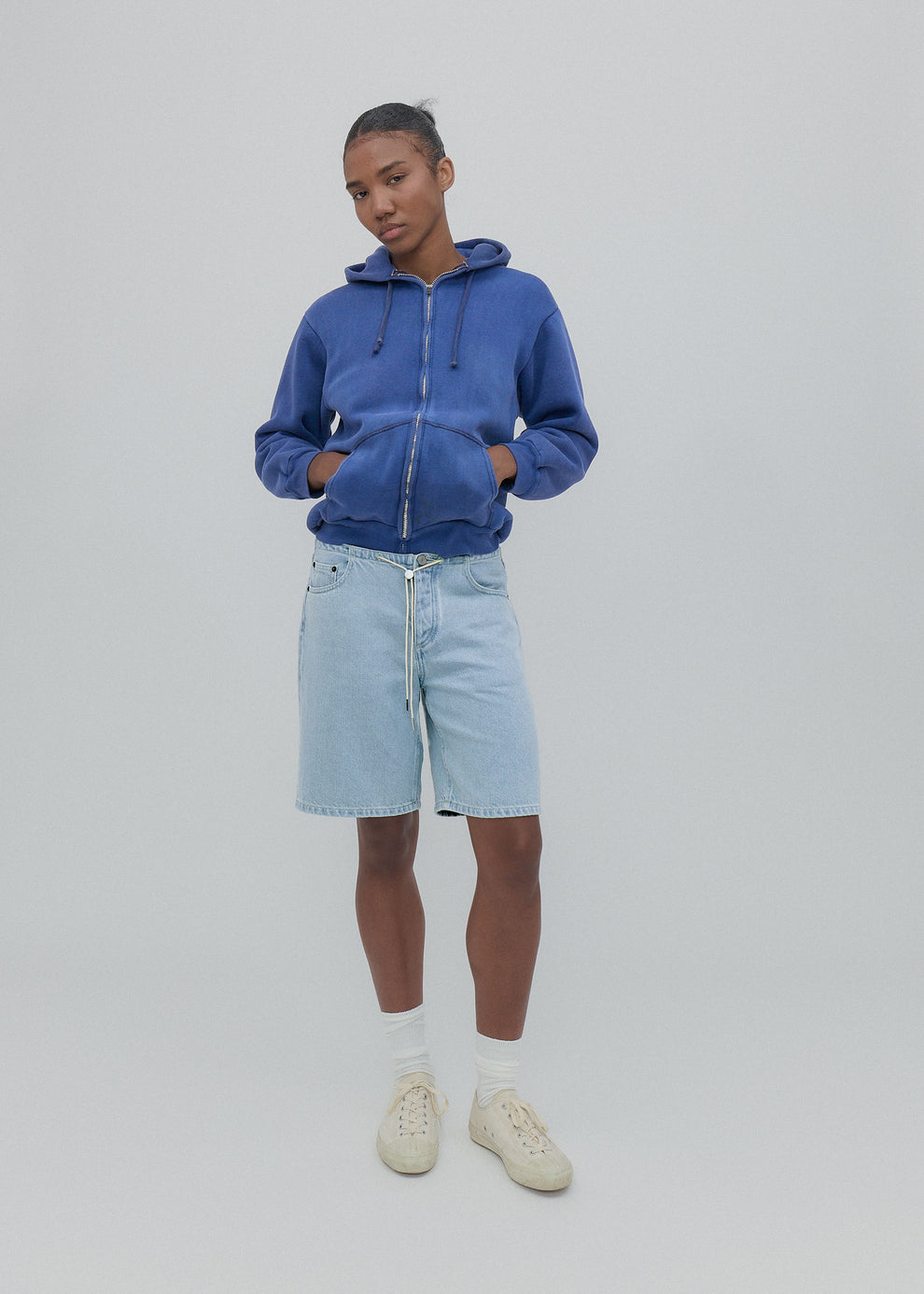 Person wearing a blue hoodie, light blue shorts, and white sneakers on a plain background