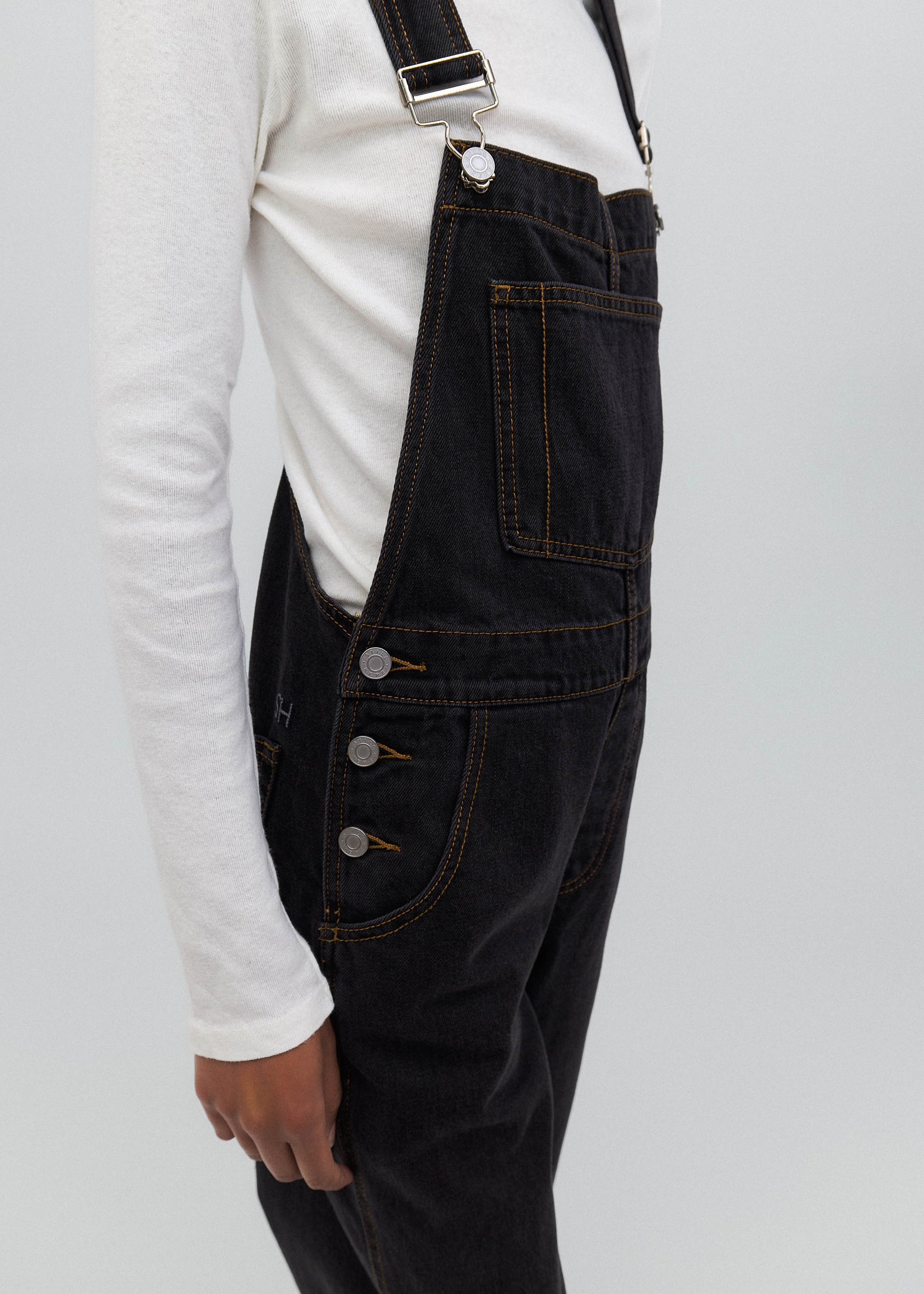 Person wearing black denim overalls with a white shirt underneath on a light gray background