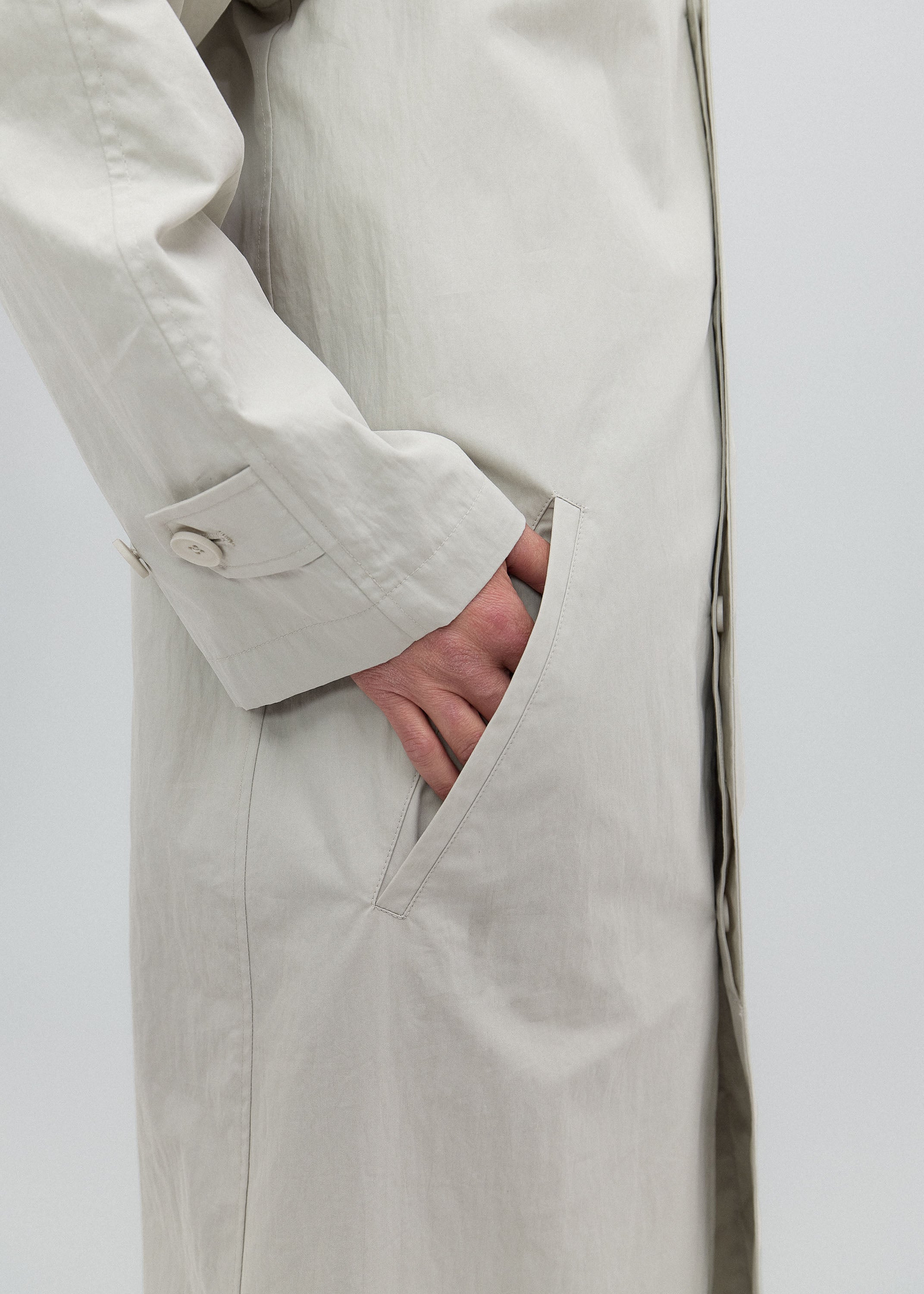 Person wearing a beige trench coat with one hand in the pocket against a white background