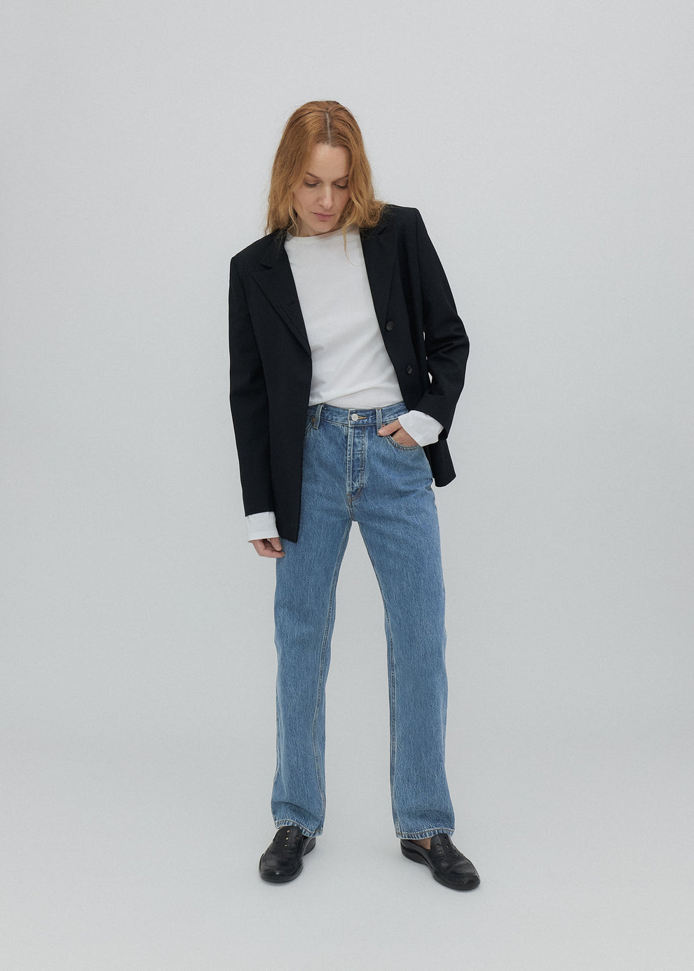 Person wearing a black blazer, white shirt, blue jeans, and black shoes on a light gray background