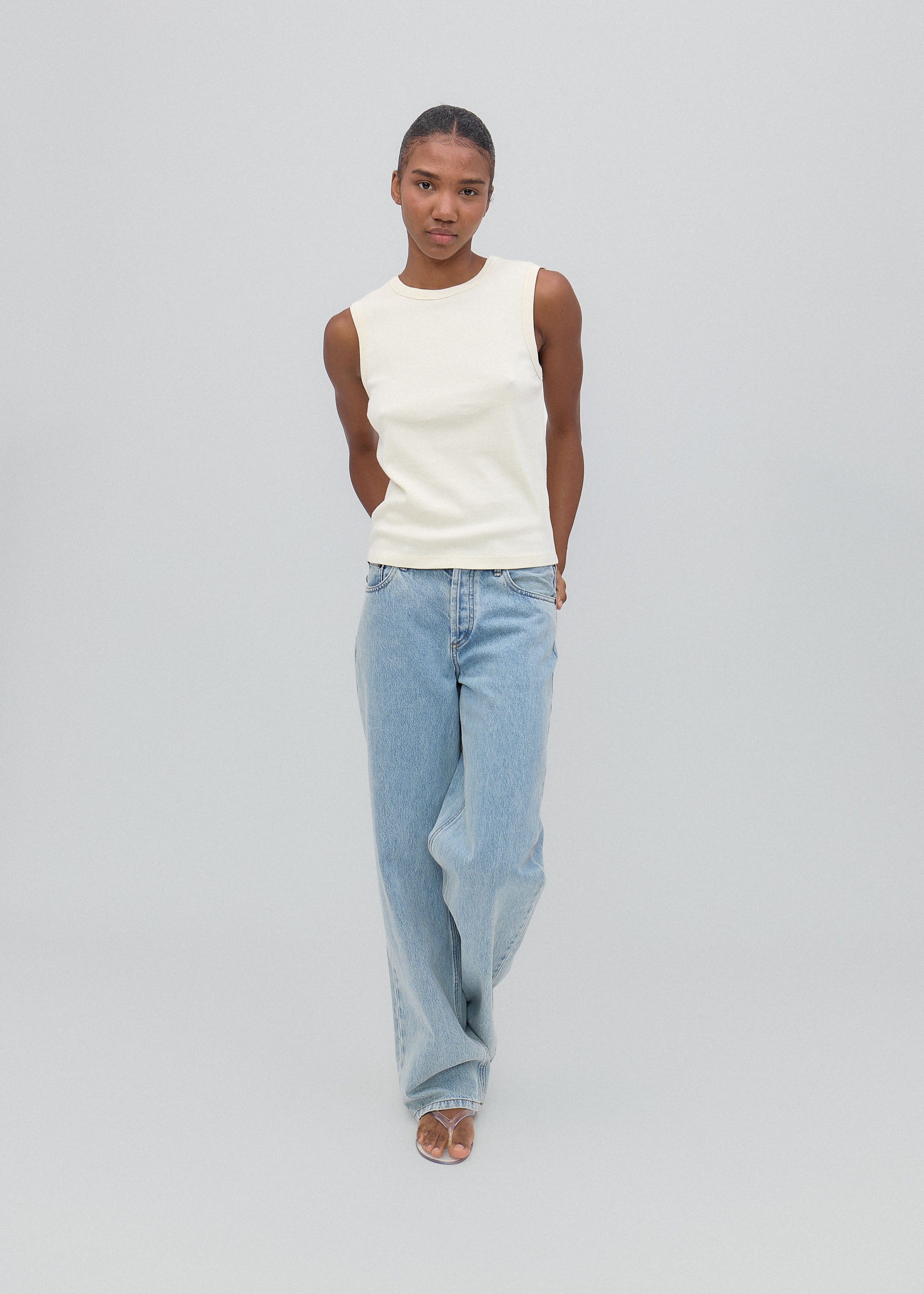 Person wearing a white sleeveless top and light blue jeans on a gray background
