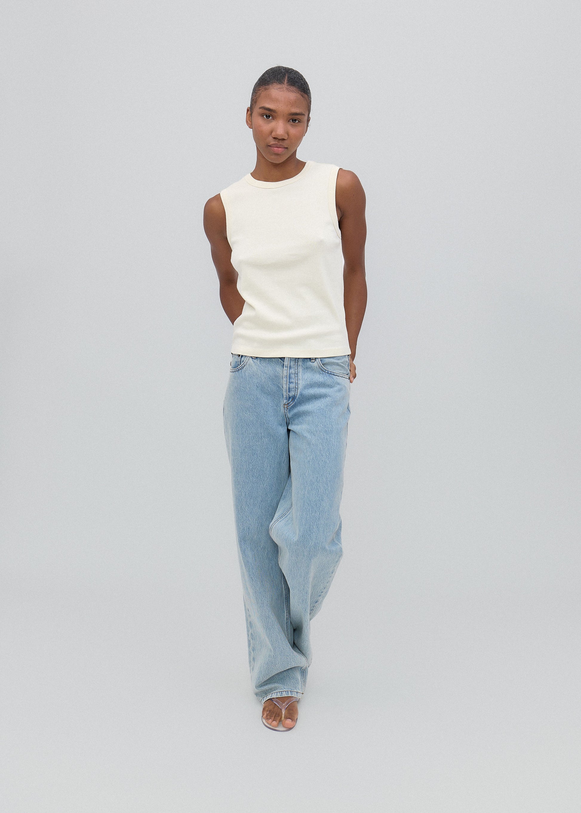 Person wearing a white sleeveless top and light blue jeans on a gray background