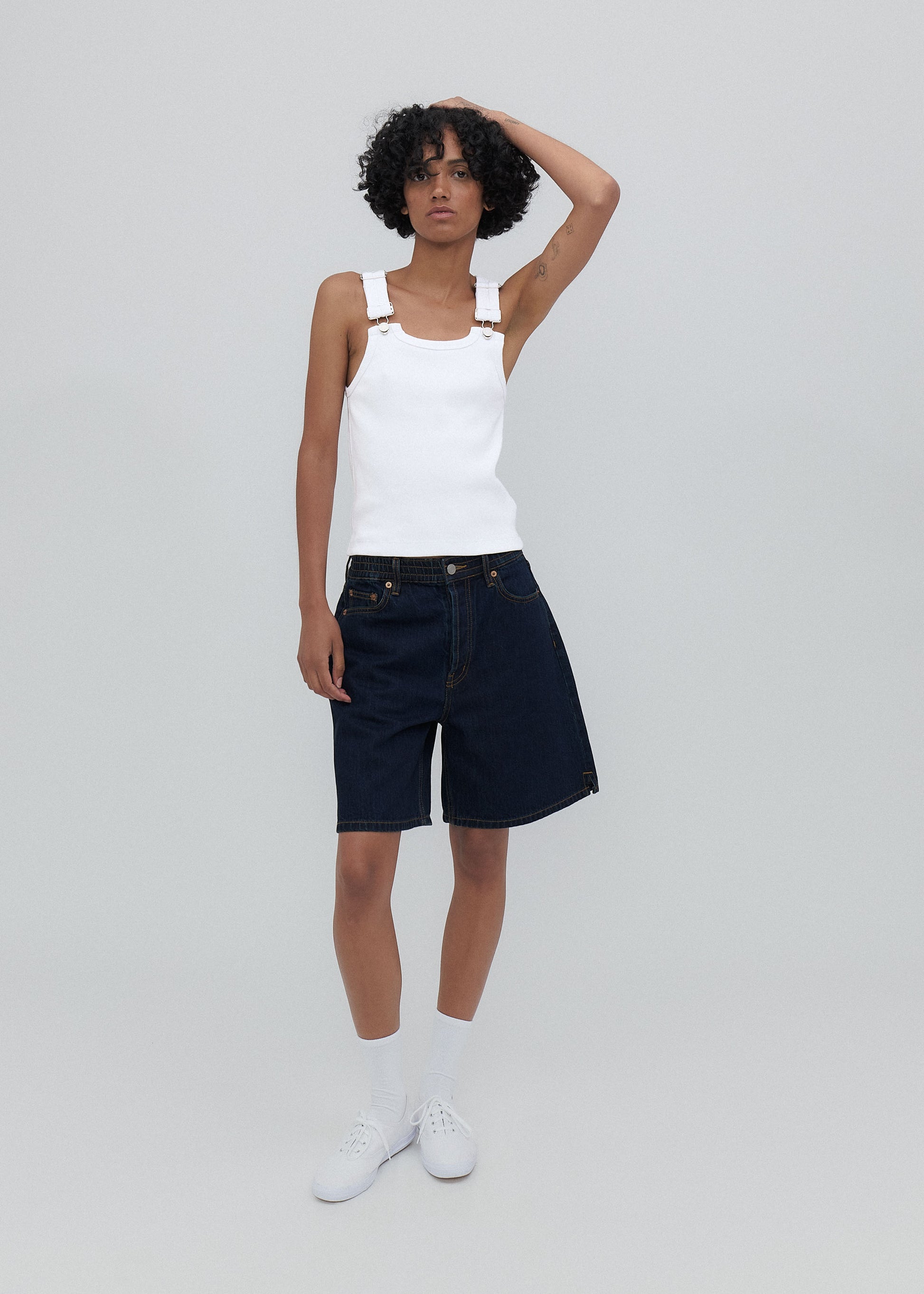 Person wearing a white tank top and dark denim shorts on a plain background