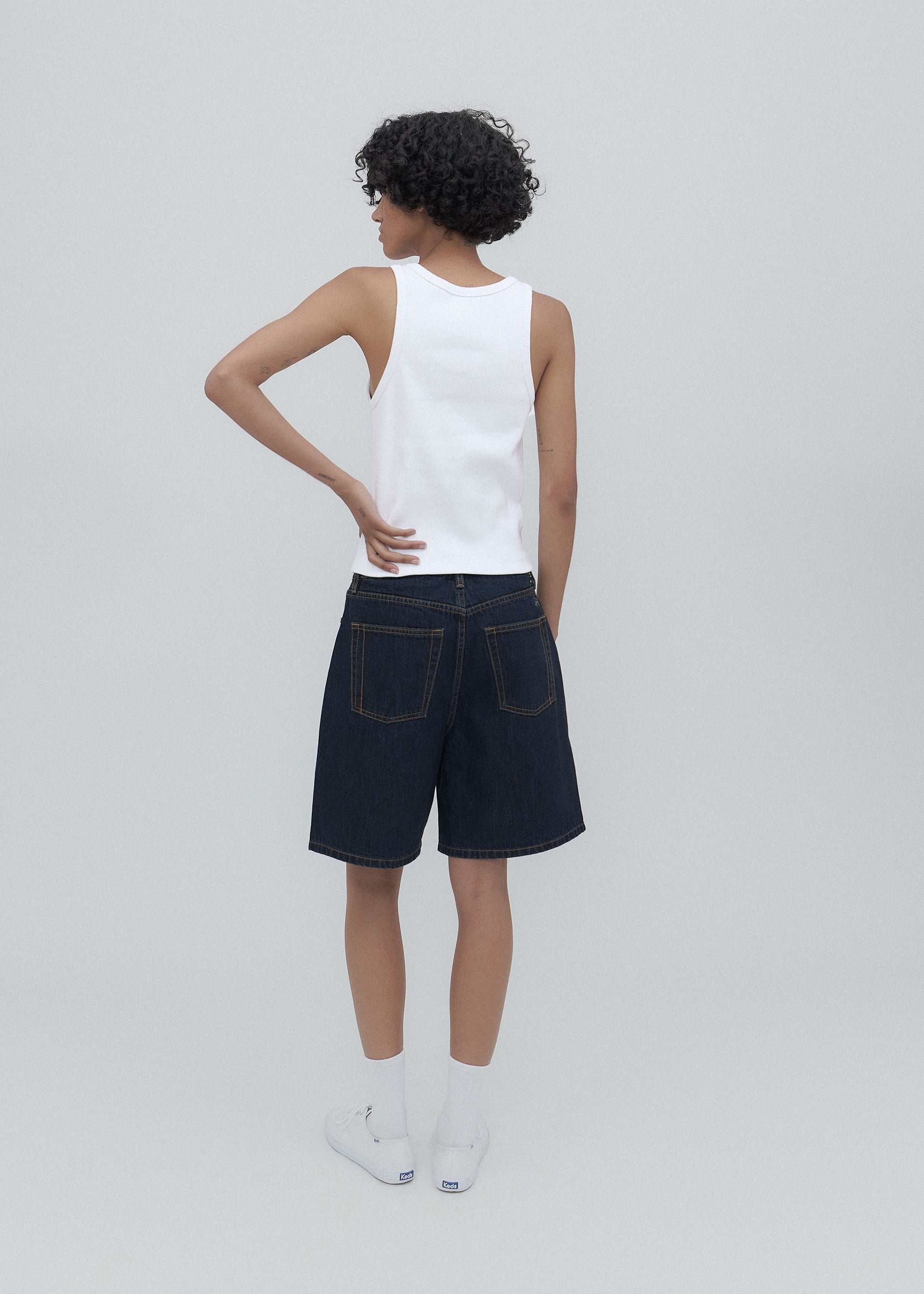 Person wearing a white sleeveless top and dark denim shorts on a light gray background
