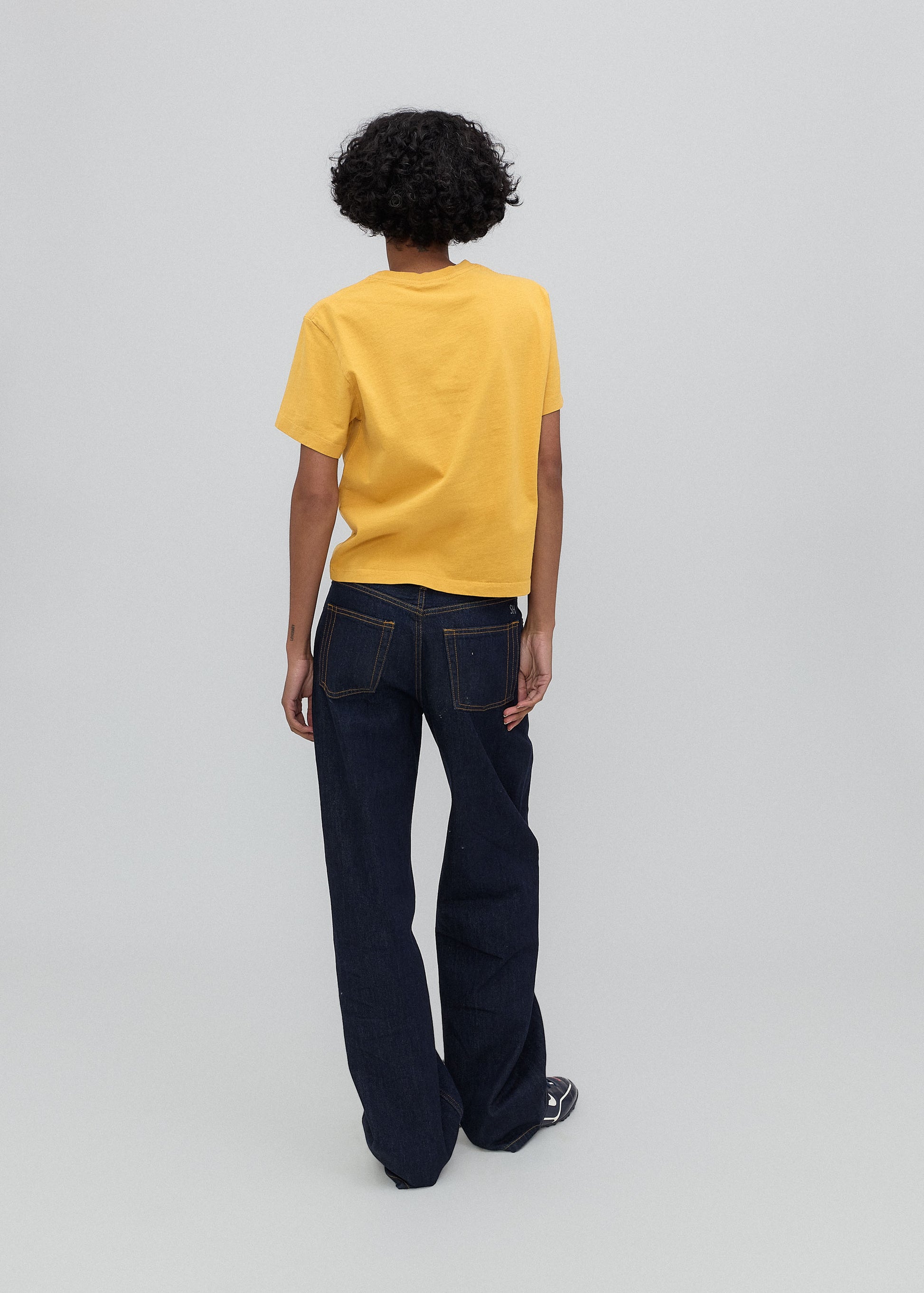 Person wearing a yellow t-shirt and dark blue jeans on a light gray background