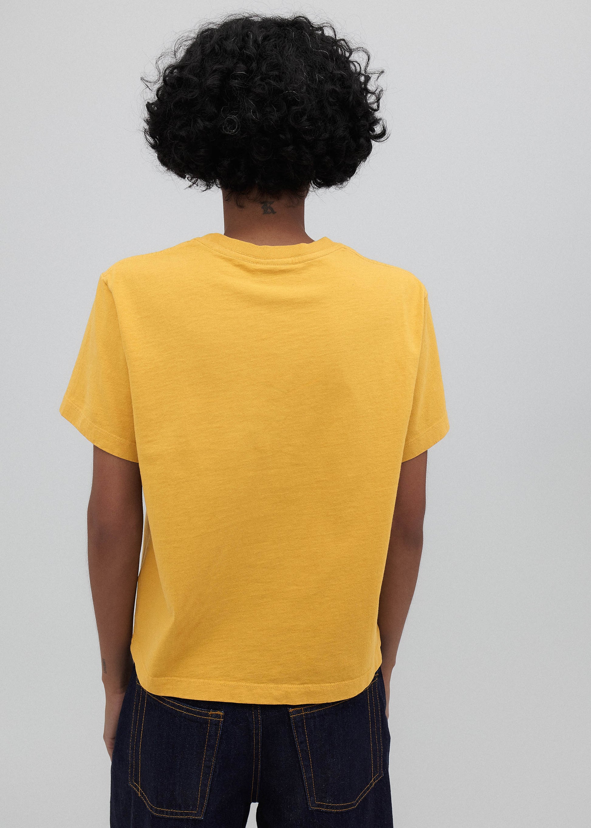 Person wearing a yellow t-shirt and dark jeans against a plain background
