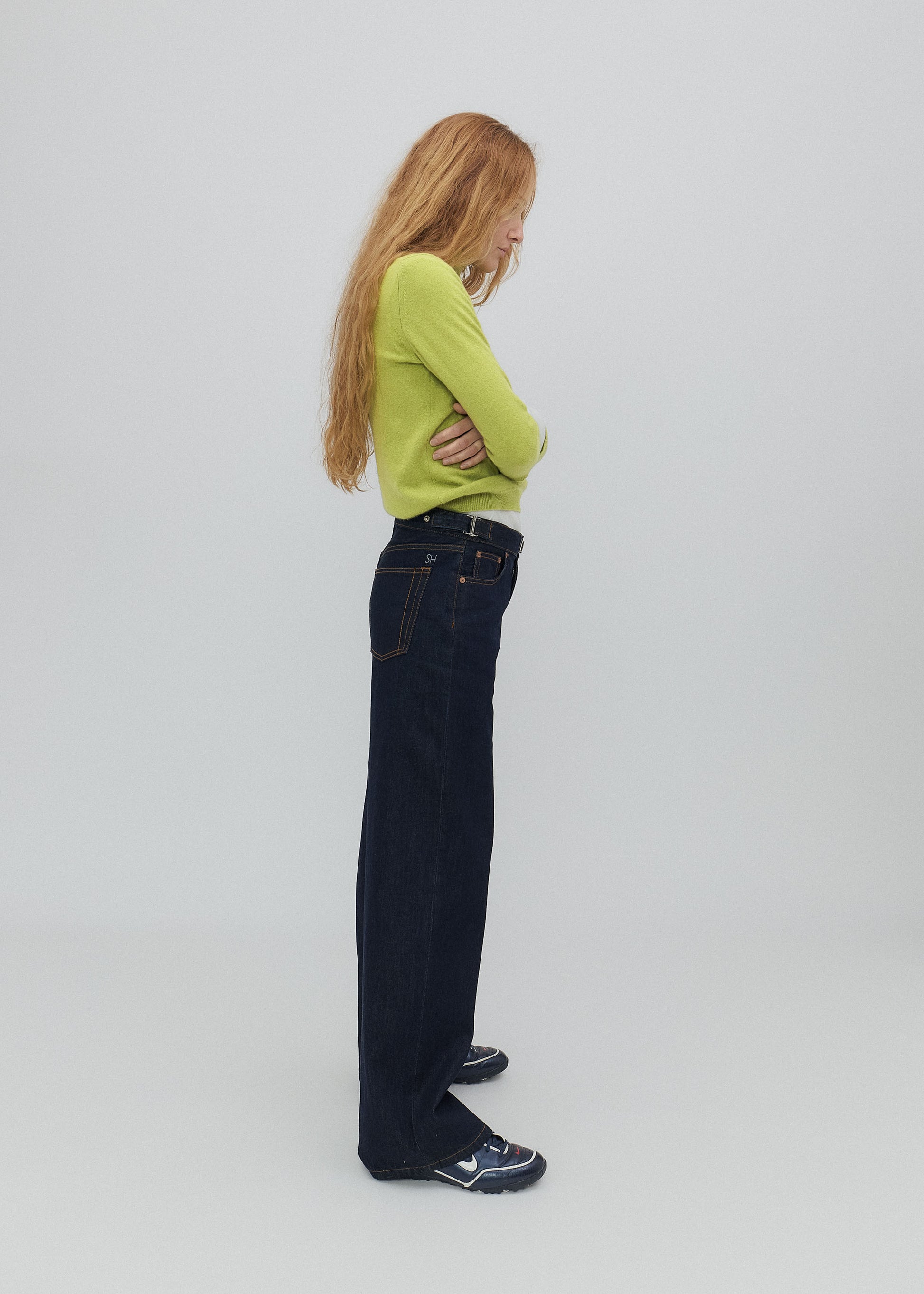 Person wearing a green sweater and dark jeans standing against a plain background