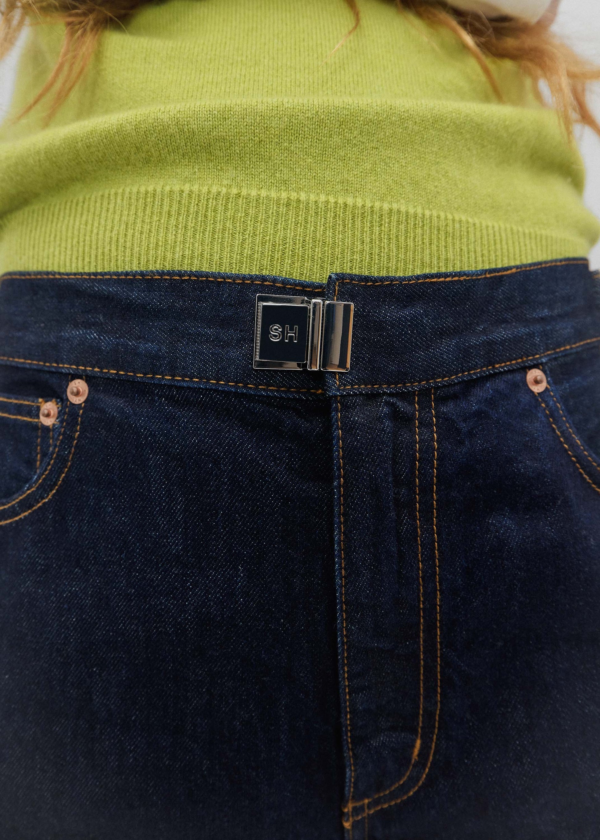 Close-up of a person wearing a green sweater and dark blue jeans with a brand logo on the pocket.