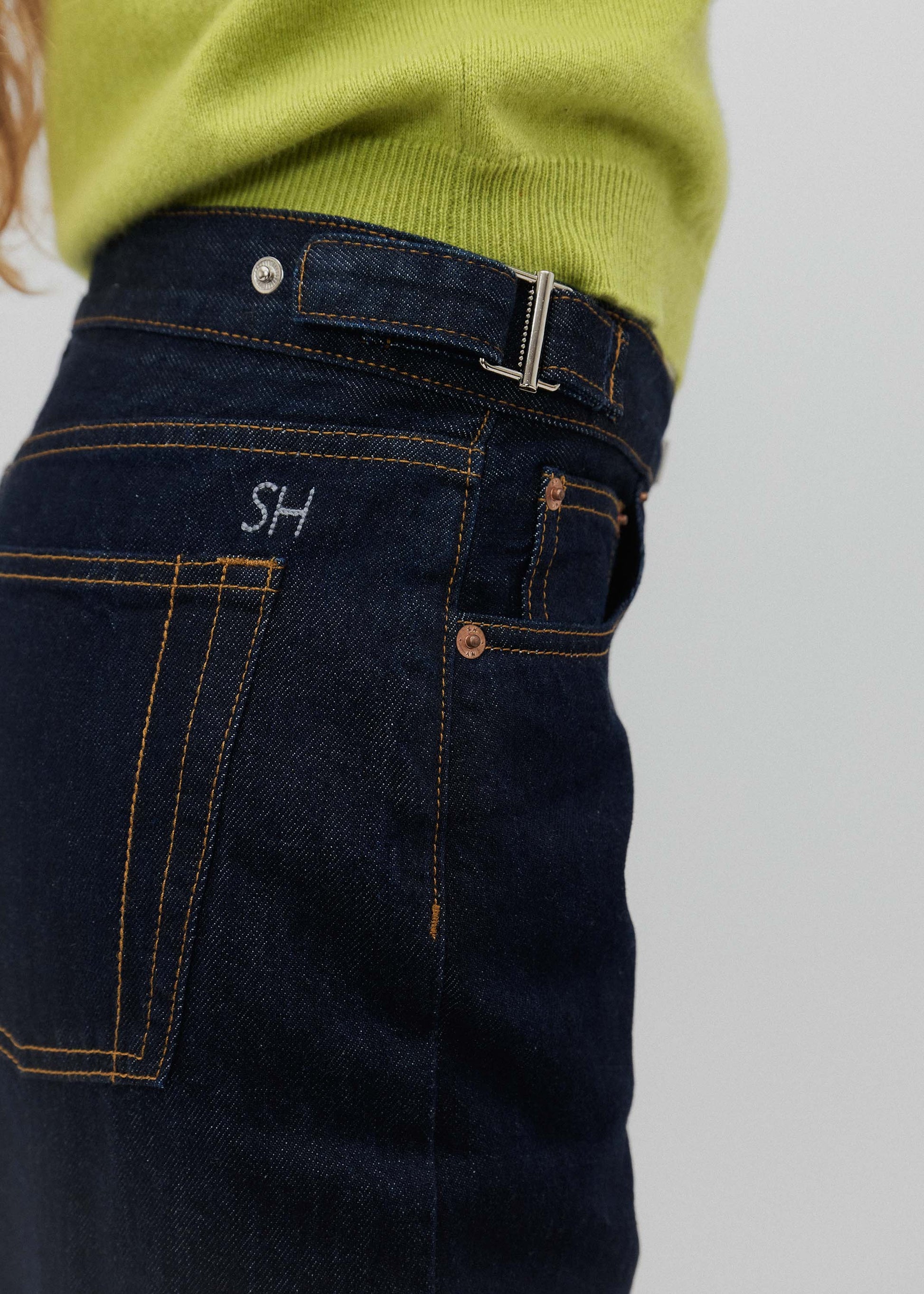Close-up of dark blue jeans with 'SH' initials on a light gray background