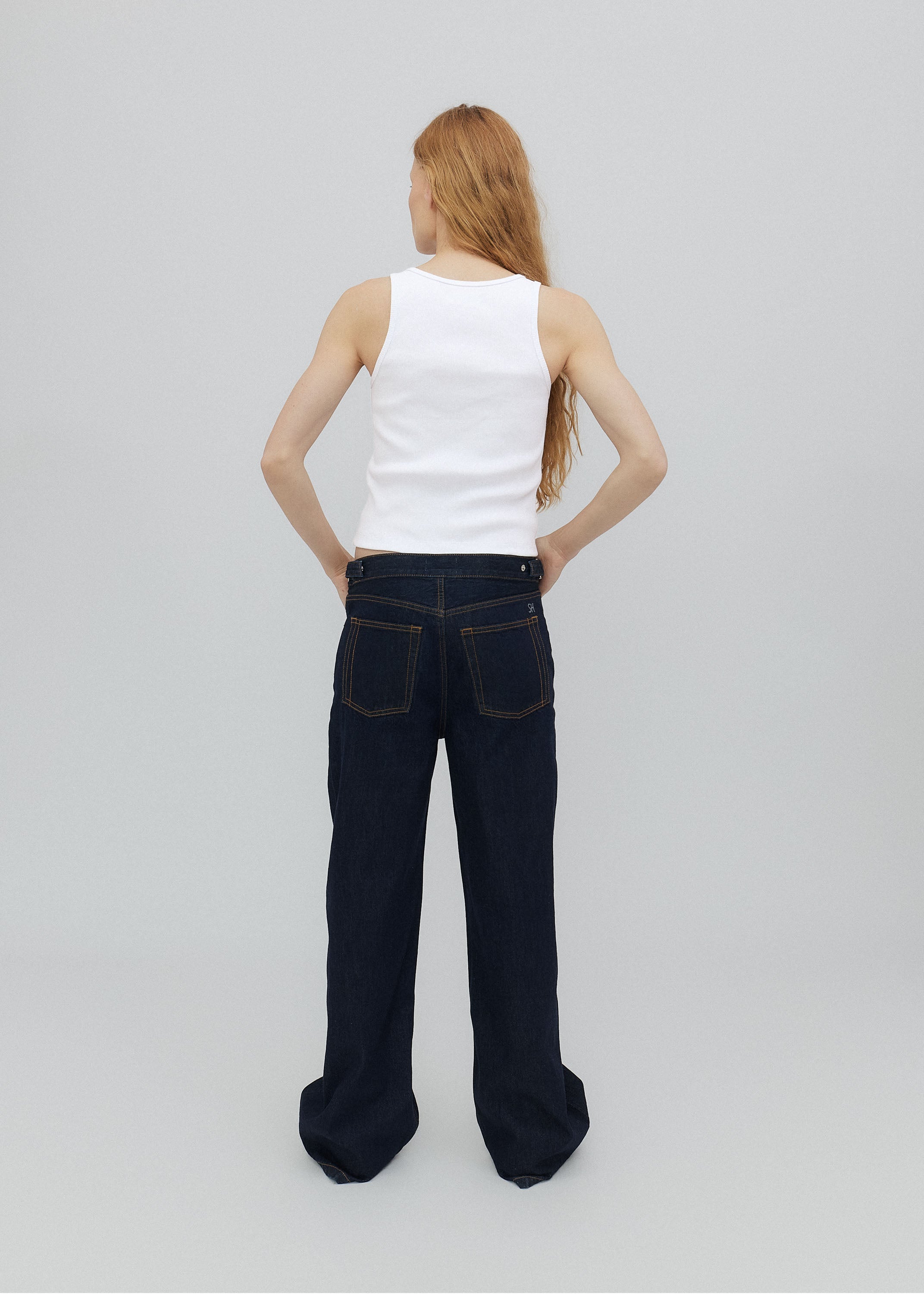 Person wearing a white tank top and dark blue jeans on a light gray background
