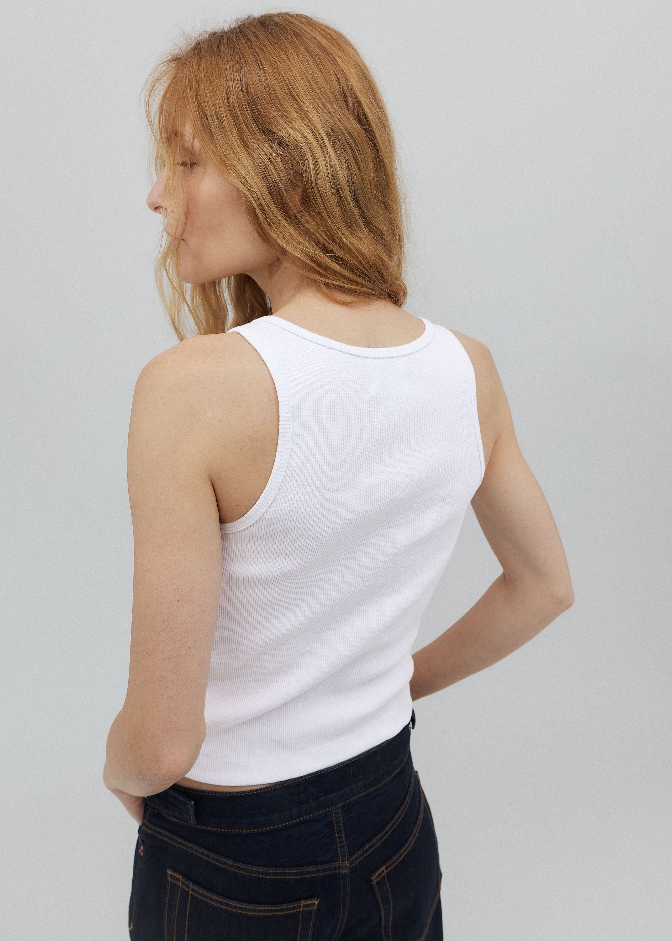 Woman wearing a white tank top and dark pants against a plain background