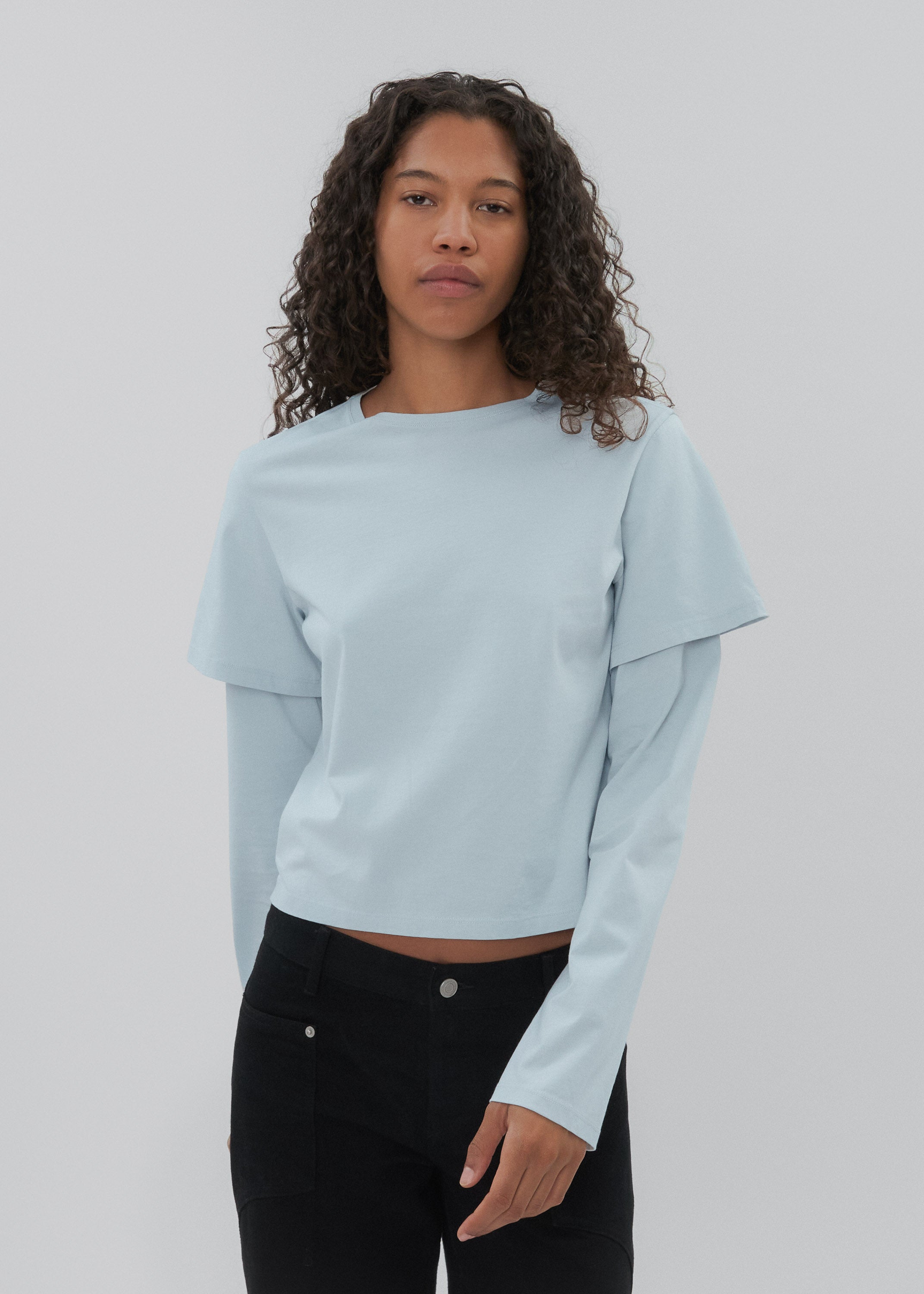 Woman wearing a light blue long-sleeve top and black pants on a plain background