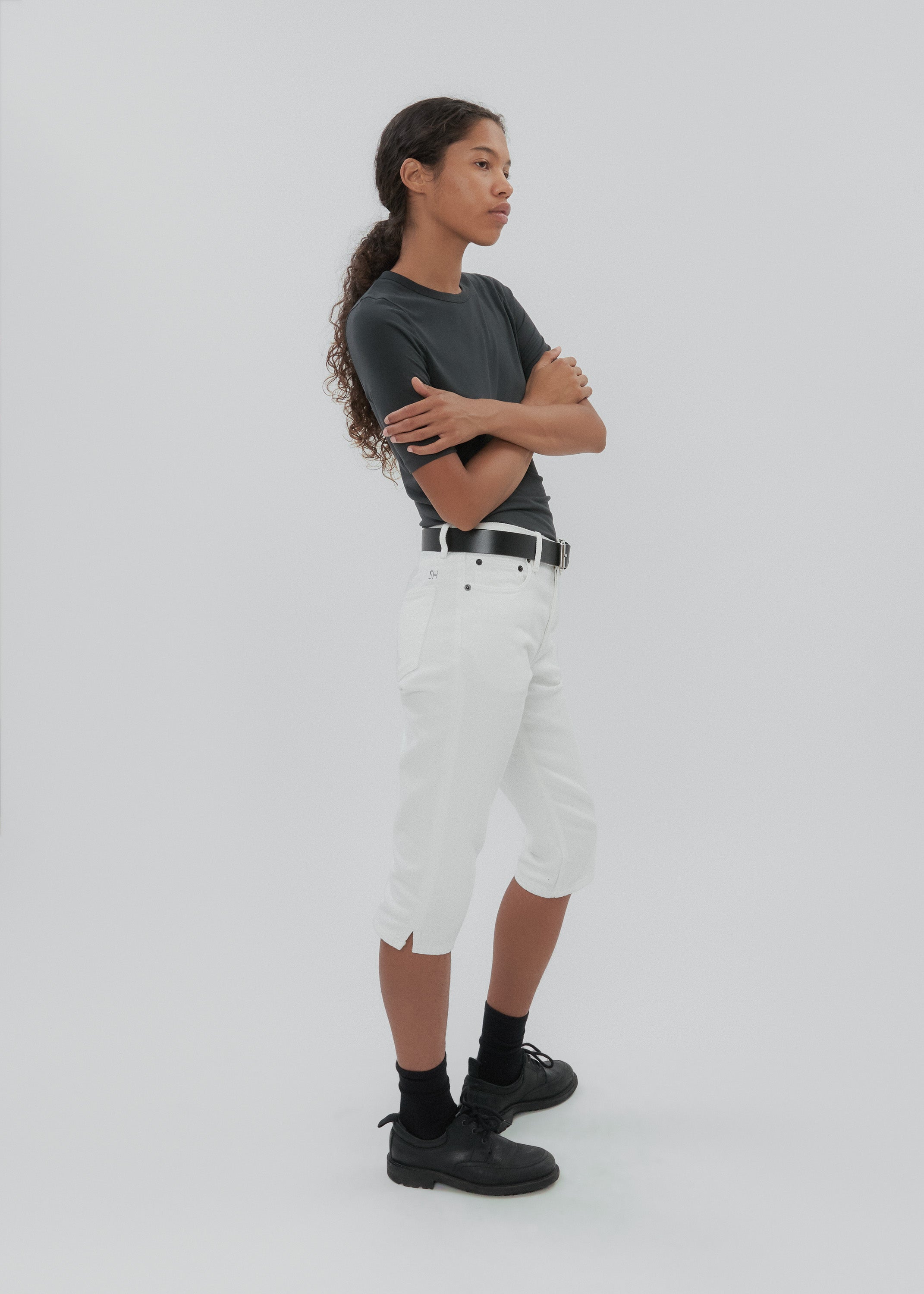 Person wearing a dark gray t-shirt, white pants, and black shoes on a light gray background