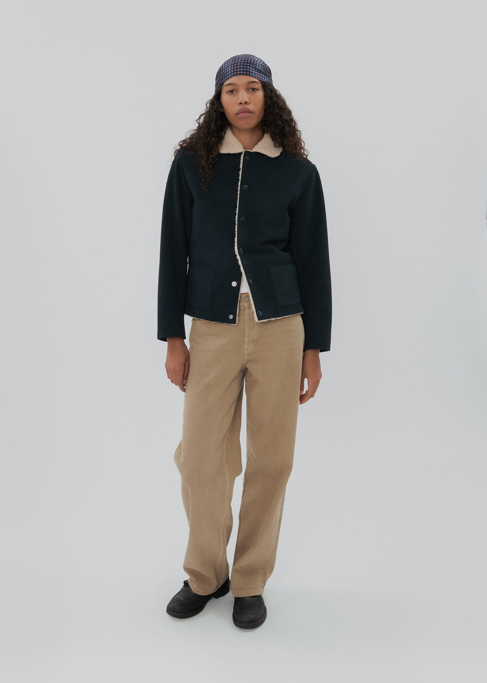 Person wearing a dark jacket, beige pants, and a patterned beanie on a white background