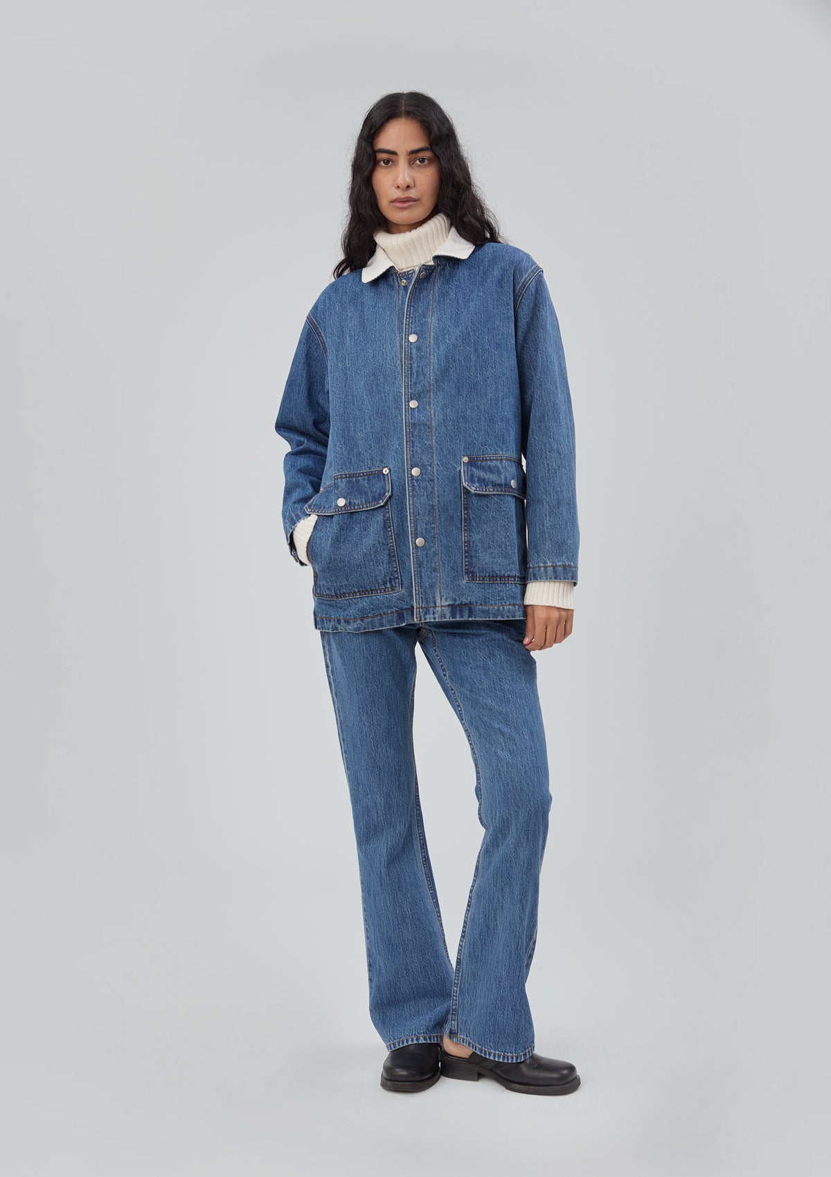 A model wearing a blue cotton Barn Jacket in Classic Blue with a white collar over a white turtleneck sweater and matching blue jeans.