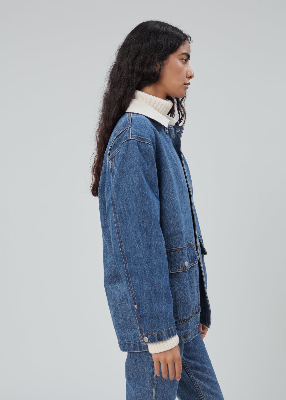 A profile view of a person wearing a Barn Jacket in Classic Blue with a white collar over a cream turtleneck sweater and matching denim jeans.