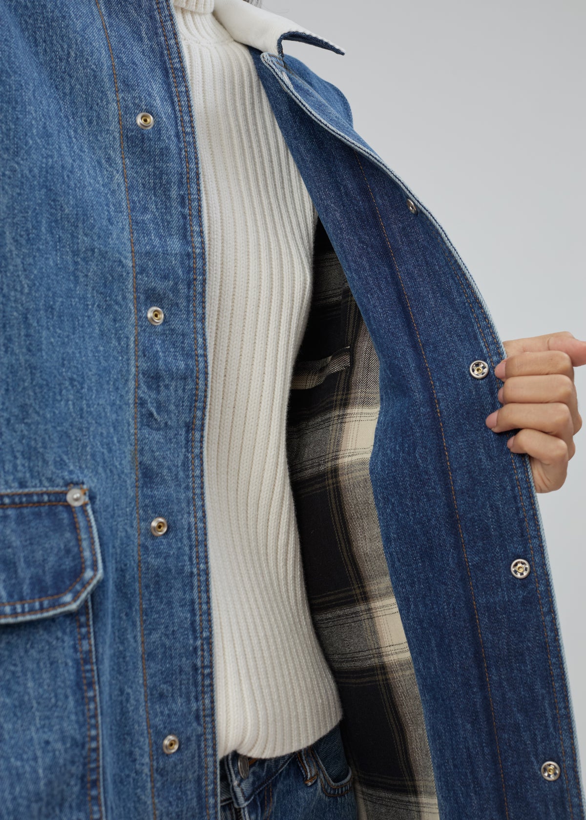 Barn Jacket in Classic Blue made of cotton denim with metal snap buttons, worn over a white ribbed sweater and showing a plaid lining.